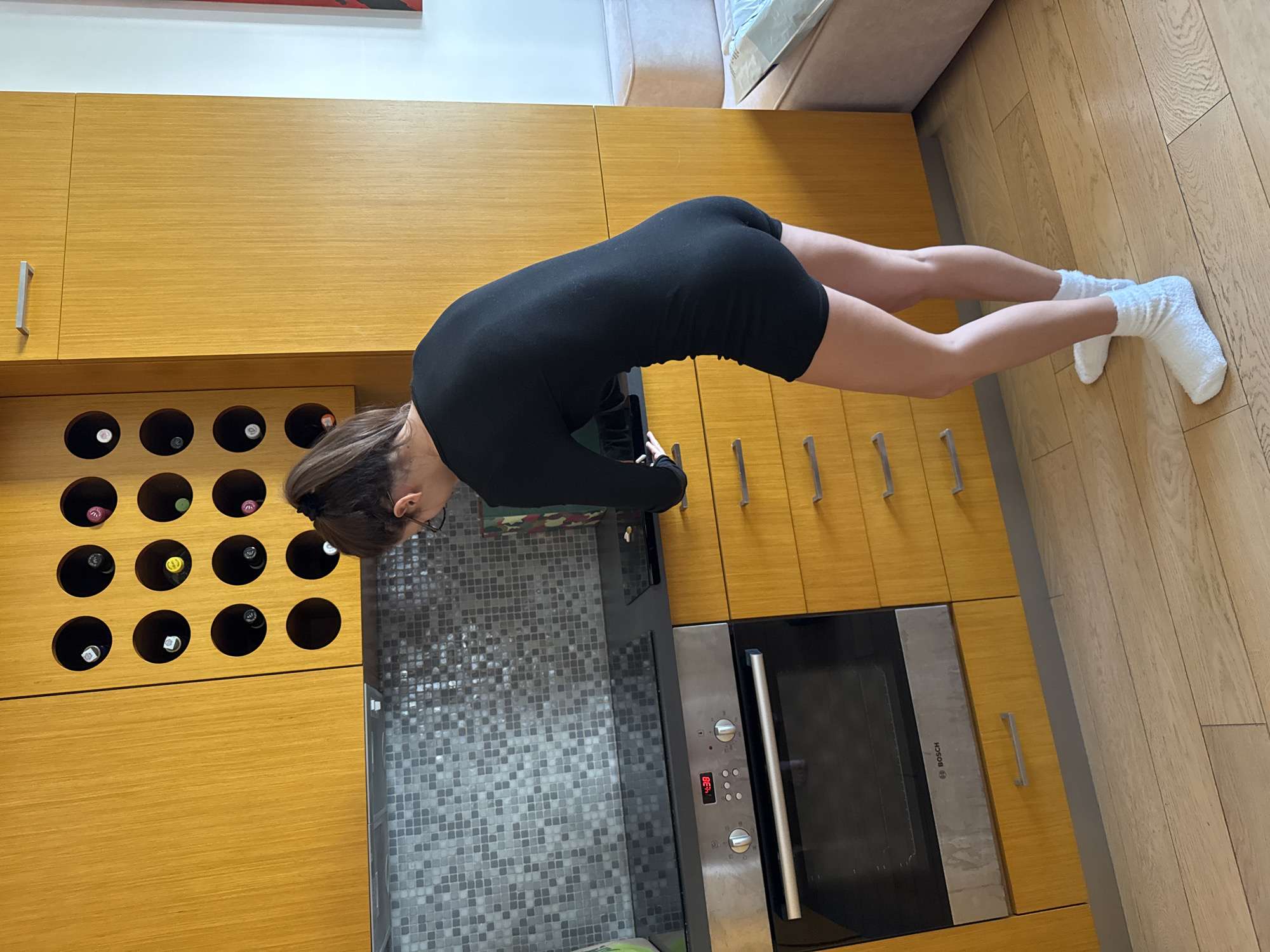 The image captures an indoor scene set within a modern kitchen, likely in a residence in Warsaw, Poland. A woman, with dark hair tied back and wearing glasses, a black long-sleeved romper or jumpsuit, and white fuzzy socks, is centrally positioned, leaning forward over a kitchen counter. Her head is bowed, suggesting she is focused on or interacting with something on the grey mosaic-tiled surface. A small, possibly green-patterned object is visible on the counter near her hands.
The kitchen is equipped with light wooden cabinetry, featuring silver drawer and cabinet handles. On the left, a built-in wine rack showcases multiple wine bottles in its circular slots. Below the counter, a built-in Bosch oven displays "4:38" on its digital clock. The floor is made of light-colored wood planks, and a light-colored sofa or upholstered furniture item is partially visible in the upper right background. The natural light filtering into the room suggests it is daytime. The woman's posture is one of absorbed attention, though her facial expression is obscured.