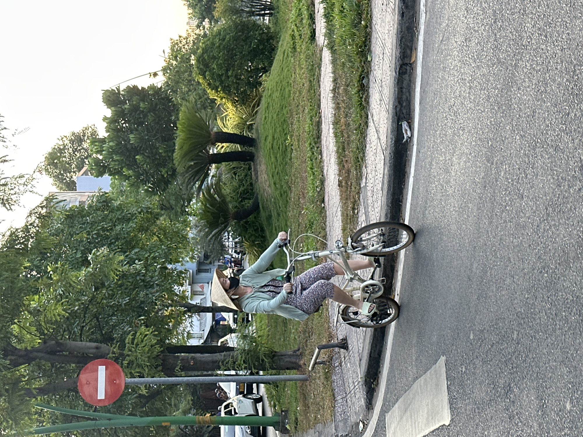 The image shows a woman riding a bicycle in Thủ Đức, Vietnam. She is wearing a conical hat, a face mask, a light green jacket, and floral print pants. She is riding on the sidewalk next to a road. To the left of the woman is a green space with trees and a red and white "Do Not Enter" traffic sign. Several cars are parked on the left side of the road. The sky is overcast. The bicycle is silver and has a basket on the front. The sidewalk is patterned with light-colored tiles.