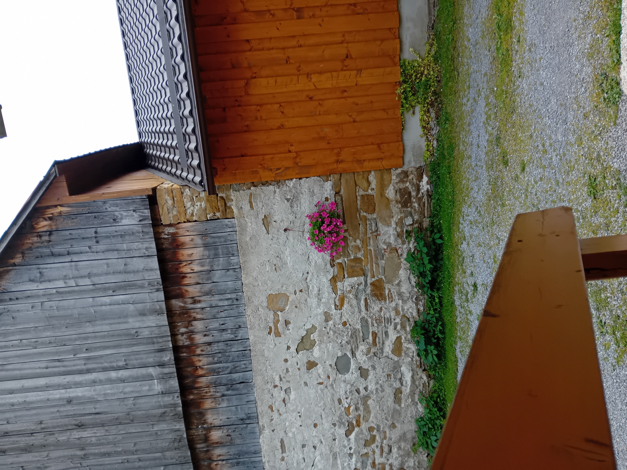 The image depicts a rustic building with a combination of aged wood siding and a stone wall. A section of the building features weathered gray wooden planks, transitioning to a lower section of darker, aged wood. Adjacent to this is a stone wall with visible mortar and patches of plaster, some of which have chipped away to reveal the underlying stones. Hanging from the stone wall is a pot of vibrant pink flowers. To the right of the stone wall, the building is clad in warm, orange-toned wooden planks. A dark, wavy-patterned roof is visible, along with a gutter. Green foliage grows along the base of the building, both in front of the stone wall and climbing up the lower portion of the orange wooden section. The ground in front of the building is covered in gravel with scattered patches of grass and small weeds. In the foreground, a blurred wooden structure, possibly a bench or railing, is visible. The sky is overcast, suggesting a cloudy day. The scene appears to be set in a rural or village environment, likely in Zuberec, Slovakia, given the architectural style and natural surroundings often found in such locations. There are no people visible in the image.