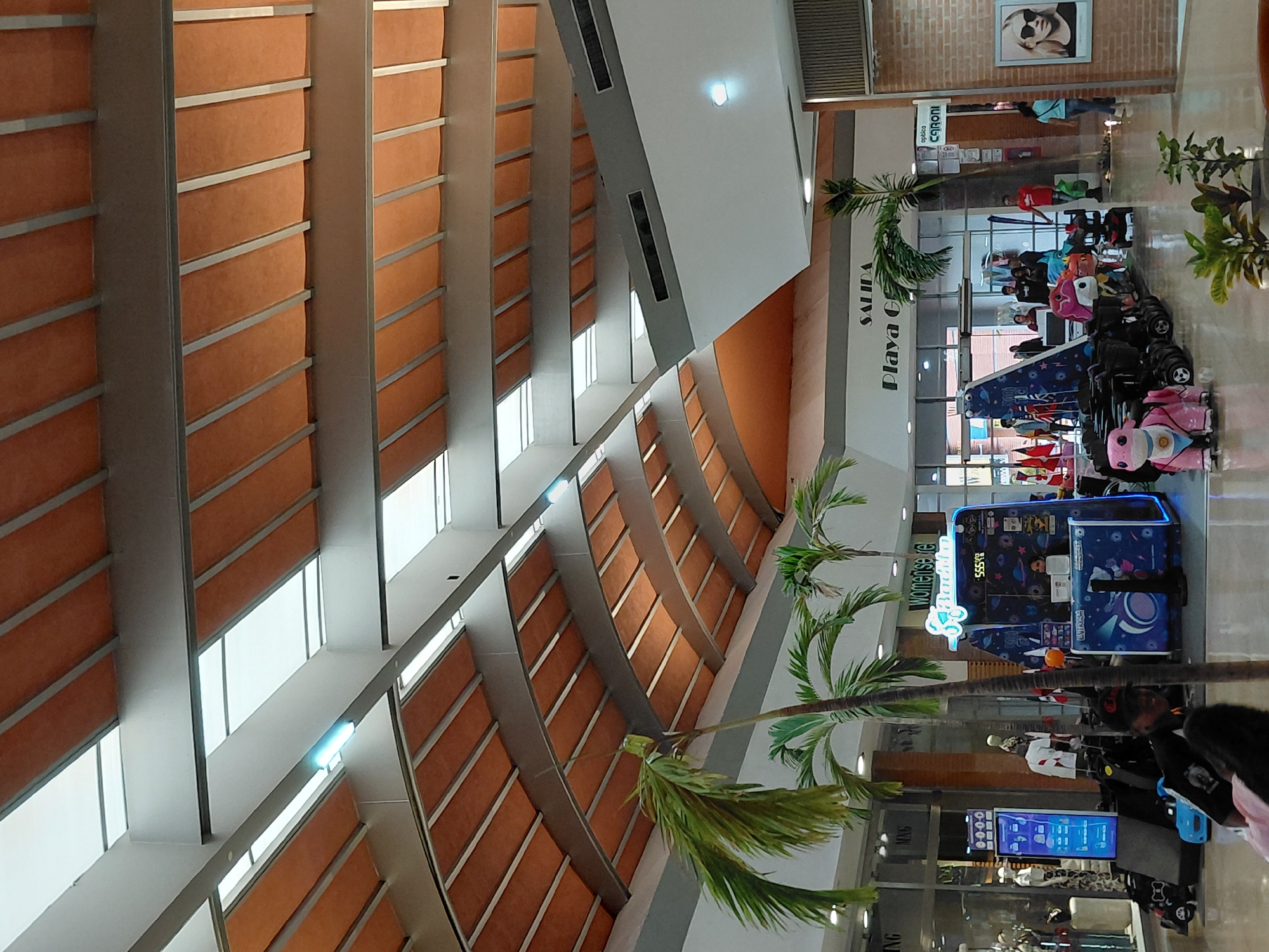 The image depicts the interior of a shopping mall, likely in Pampatar, Venezuela, featuring an arched ceiling with translucent panels and orange fabric. Palm trees are visible throughout the space. In the foreground, a child's ride is parked, along with a kiosk offering a game. People are seen browsing shops and walking through the mall. Signage includes "SALIDA" (Exit) and "Playa G" for a store. To the right, there's an optical shop called "CARONI" and a framed portrait of a woman wearing sunglasses. The environment appears to be well-lit and spacious, typical of a modern shopping center.