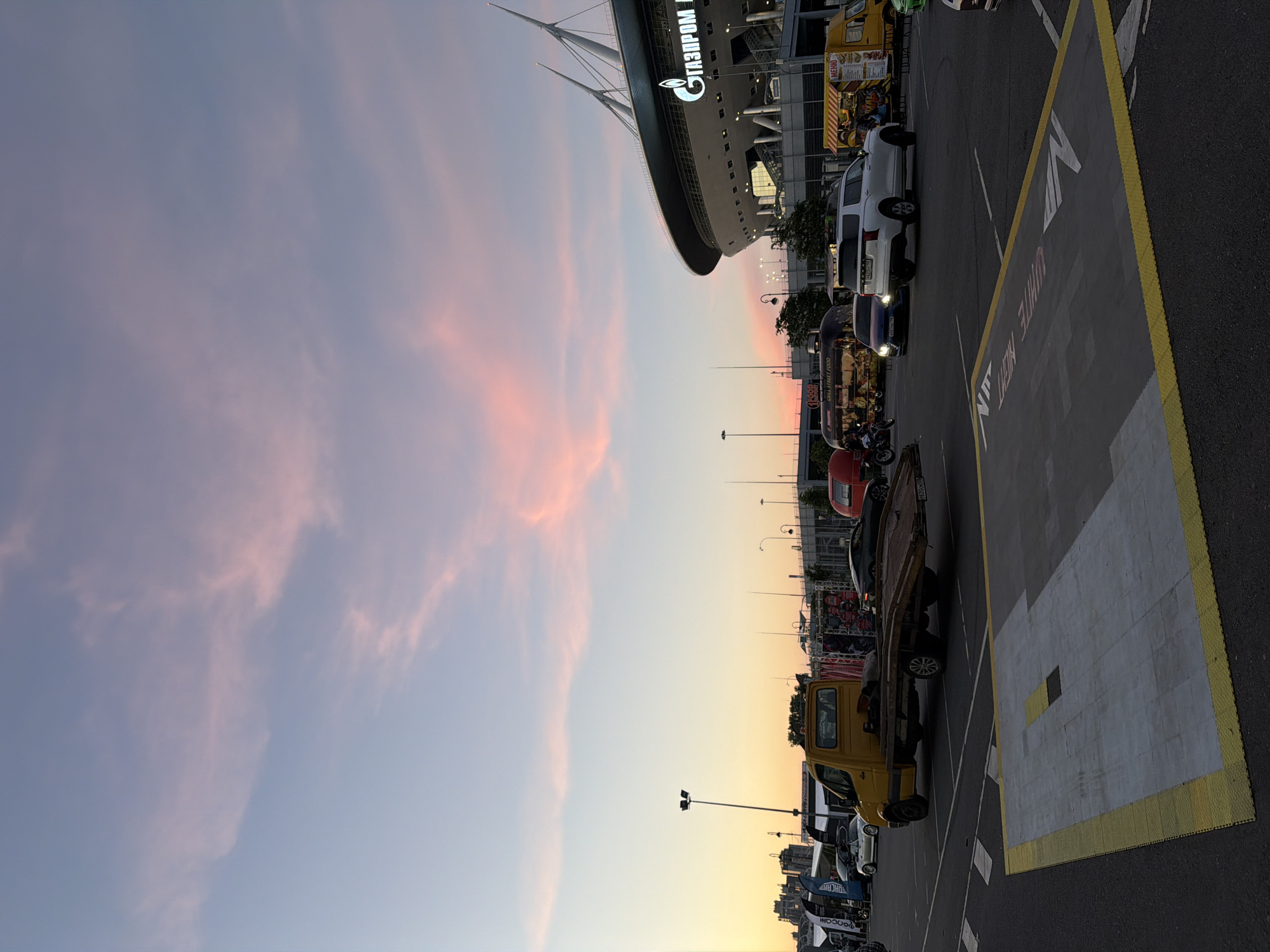 The image shows a parking lot in front of a large stadium at dusk. The sky is filled with soft pink and blue clouds. The stadium, which has the Gazprom logo on it, dominates the background. Several vehicles are parked in the lot, including a yellow tow truck, a white SUV, a blue sedan, and a motorcycle. There are also some food trucks and tents set up. The overall scene suggests a pre-event atmosphere or an event that has just concluded.