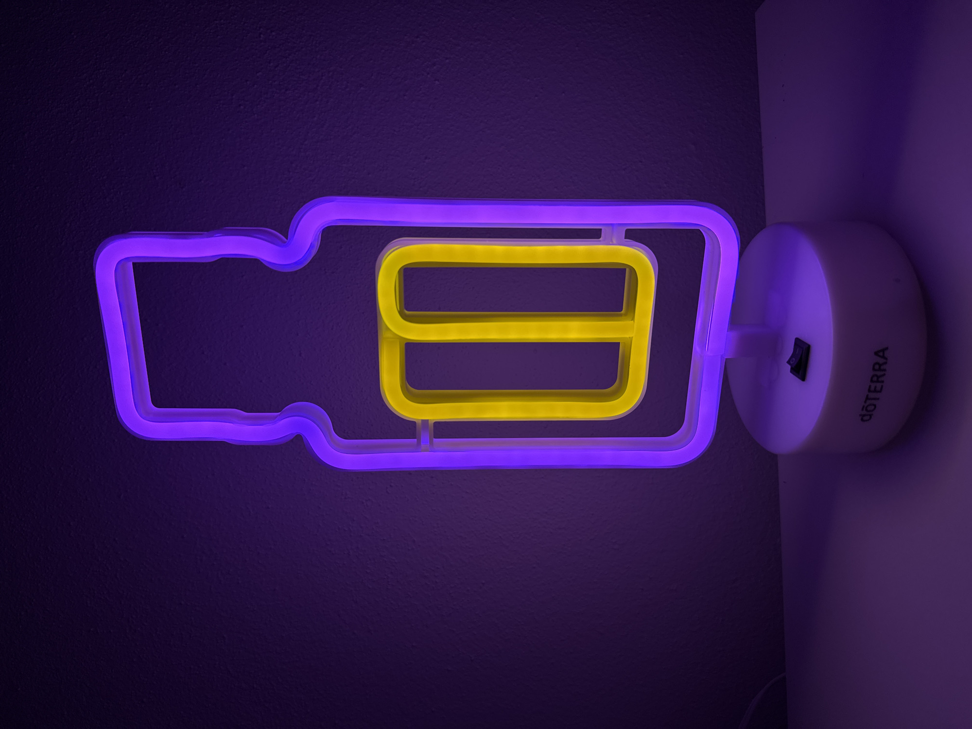 A close-up, eye-level, indoor shot features a white, circular base with a black power switch and the word "dōTERRA" printed in black on its surface. Resting on the base is a neon sign shaped like a stylized bottle. The outline of the bottle is illuminated in purple neon, and within it, a rectangular shape is illuminated in yellow neon. The background is a textured, dark purple wall, and the surface beneath the base is white.