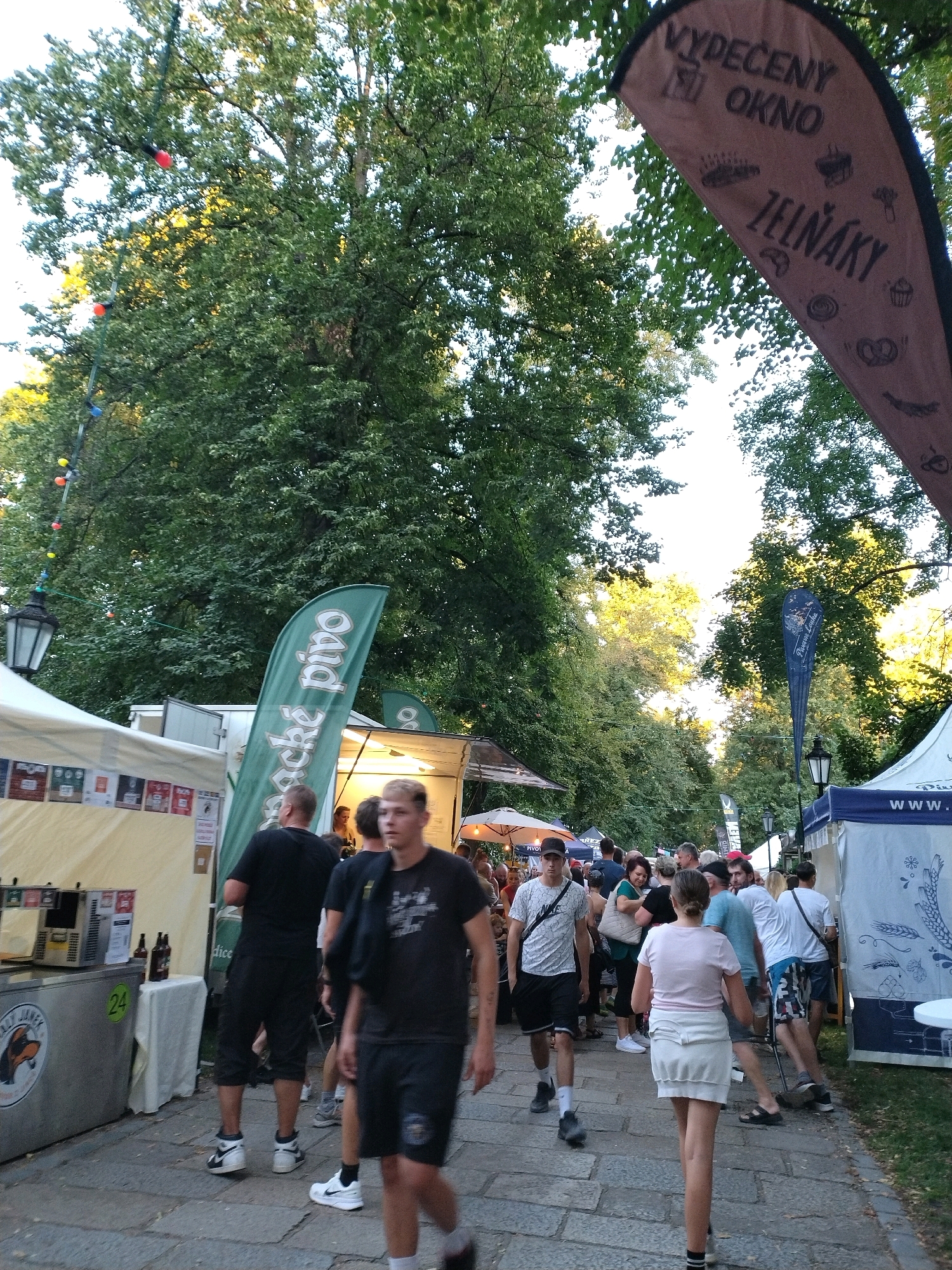 The image depicts an outdoor event or market, likely a beer festival, in Písek, Czechia, given the presence of flags with Czech text like "VYPEČENÝ OKNO ŽELŇÁKY" and "České pivo". The scene is crowded with people of various ages and genders, many of whom are looking at stalls or interacting with each other. In the foreground, a young man in a black t-shirt and shorts is walking towards the camera, with another man in a similar outfit behind him. To the left, a stall displays beer taps and bottles, with a sign featuring a dog mascot. Several flags, including a large brown one with text and illustrations of baked goods, and a green one with "České pivo" written vertically, are visible. Trees provide shade over the area, suggesting it is a warm day. The ground is paved with cobblestones. The overall atmosphere is lively and social.