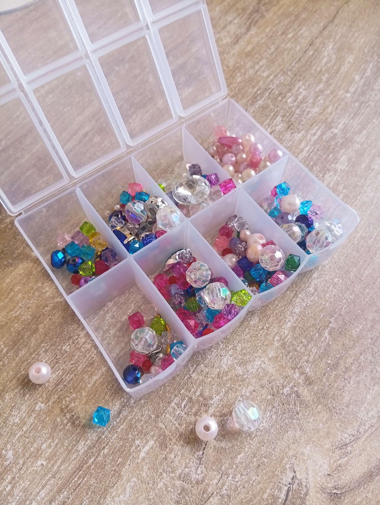 This image depicts a clear plastic bead organizer with multiple compartments, filled with various colorful beads. The organizer is open, and the beads are predominantly in shades of pink, blue, green, clear, and purple. Some of the beads are faceted and transparent, while others are opaque pearls. A few individual beads, including a pink pearl, a blue faceted bead, and a clear faceted bead, are scattered on the wooden surface next to the organizer. The background is a light brown wooden surface with visible grain. There is no indication of a specific location in Hrodna, Belarus. The scene is static and shows a collection of crafting materials.