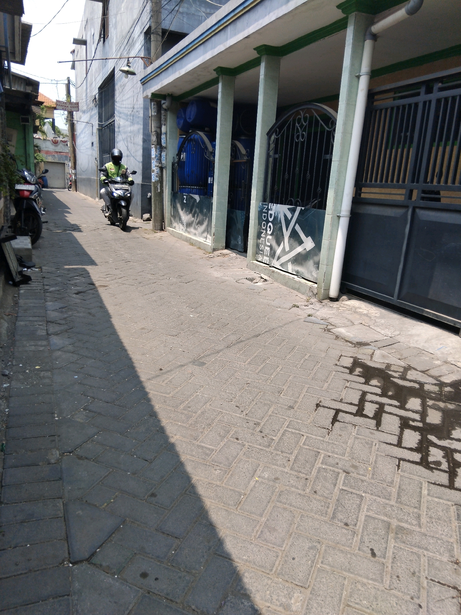 In Kepuh Kiriman, Indonesia, a motorcyclist wearing a bright green vest and a black helmet is riding down a narrow, paved alley. The alley is flanked by buildings on both sides. The building on the left appears to be a residential structure with a green facade and a corrugated metal roof. The building on the right is a more substantial building with a gray exterior, white trim, and a covered walkway supported by columns. A large black metal gate is visible in front of this building. The pavement in the alley is made of interlocking bricks. Sunlight casts a long shadow across the alley, indicating it is likely daytime. There are several parked motorcycles on the left side of the alley. The scene depicts a common urban streetscape in Indonesia, characterized by its narrow alleys, dense development, and vibrant street life.