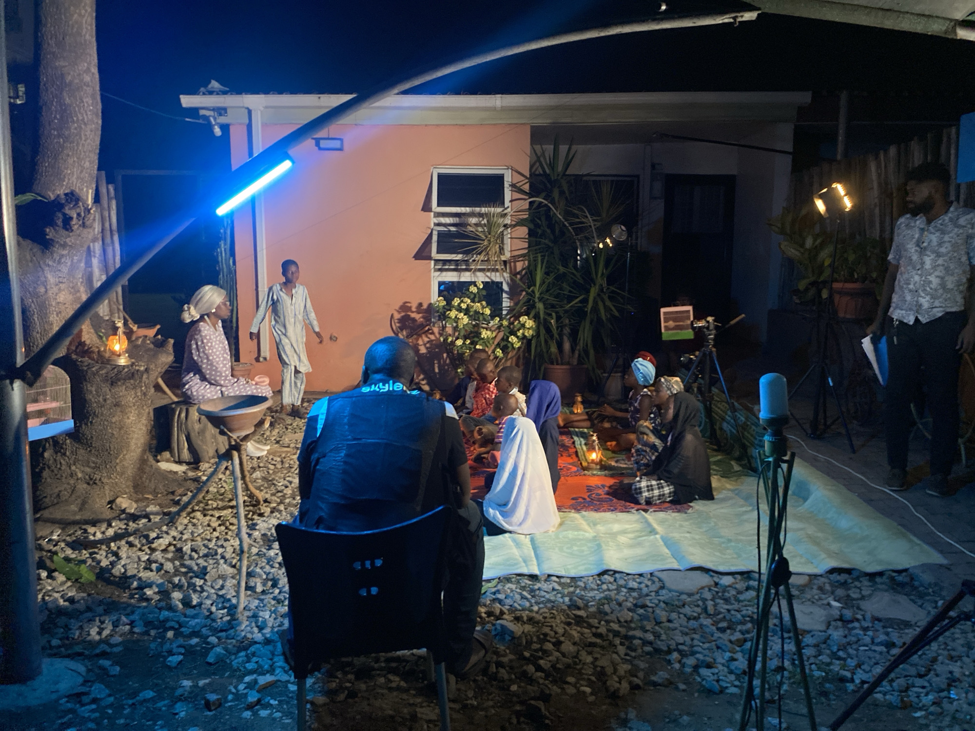 The image depicts an outdoor night scene in Barden Yana, Nigeria. A group of people are gathered on a rug placed on a gravelly ground, with a house in the background. Some individuals are seated on the rug, while others are standing nearby. A man stands on the right holding papers. Lighting equipment, including lamps and a microphone stand, are visible around the scene. A tree is on the left. The house has an orange facade with visible windows. The overall atmosphere is one of a film or stage production.