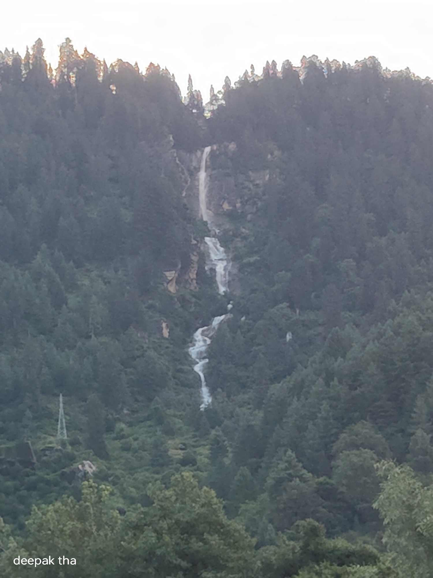The image depicts a lush, mountainous landscape with a prominent waterfall cascading down a rocky cliff face. The waterfall appears to be multi-tiered, with water flowing in streaks and rivulets. The slopes are densely covered with dark green coniferous trees, creating a textured, natural environment. In the lower left portion of the image, a metal electricity pylon stands against the backdrop of trees. The sky above is a bright, washed-out white, suggesting either dawn or dusk. The overall impression is one of a remote, natural setting, likely in a mountainous region. The text "deepak tha" is overlaid on the bottom left corner of the image. Based on the context of Vashisht, India, this scene is likely a view of the natural beauty surrounding the town, possibly a well-known waterfall in the vicinity.
