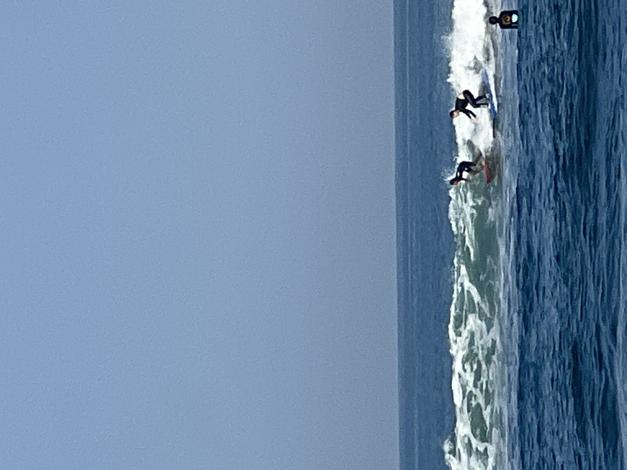 The image depicts a bright, clear day at the ocean in North Kuta, Indonesia, with a large expanse of calm blue water dominating the foreground. In the middle ground, a moderate wave is breaking, creating white foam and spray. Three individuals are visible in the water. Two surfers are actively riding the breaking wave. The surfer on the left is on a red surfboard, wearing a black wetsuit. The surfer on the right is on a blue surfboard, also wearing a black wetsuit. Further to the right and slightly further out in the water, a third individual, appearing to be wearing a dark wetsuit, is present. The background consists of a vast, clear, light blue sky with no clouds. The horizon line between the ocean and the sky is sharp and distinct.