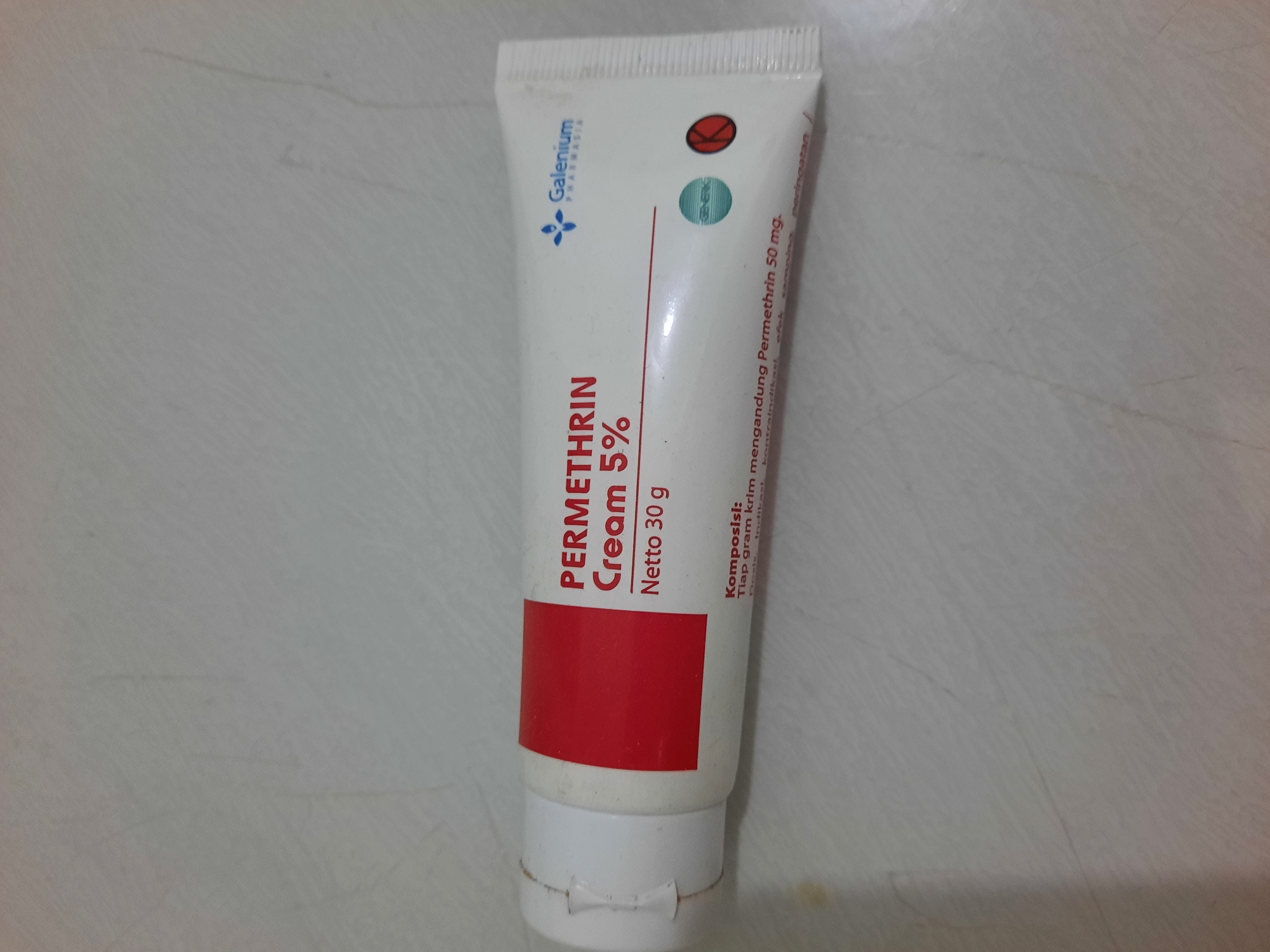A white tube of "PERMETHRIN CREAM 5%" with a red band is placed horizontally on a light-colored, textured surface, likely a table or countertop. The tube has text in Indonesian and English, including the brand name "Galenium Pharmasia," product name, net weight, and composition. There are also circular icons indicating "Generik" and a red circle with a white "K." The lighting suggests an indoor environment. The location in Lhokseumawe, Indonesia, is not visually represented in the image itself, but can be inferred from external information.