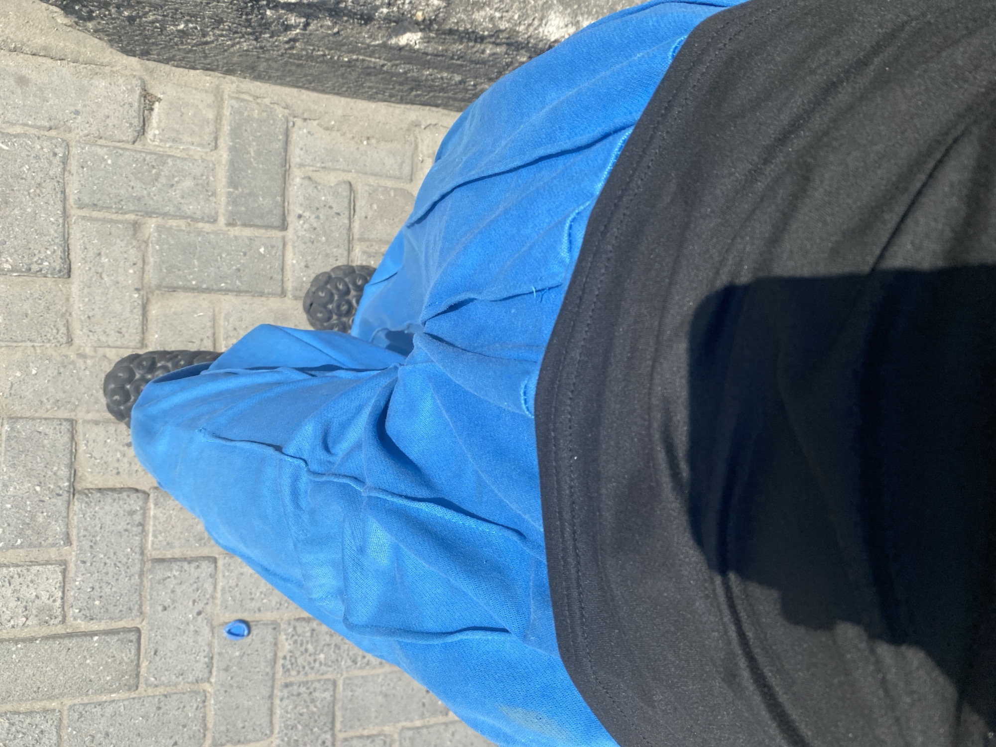 A street-level view shows a person standing on a gray brick sidewalk. The person is wearing blue pants with a textured, layered design, and a black shirt. The person's feet are clad in dark-colored sandals. A small, blue object lies on the sidewalk near the pants. The background is a concrete brick surface. The location is the city of Iranla, Nigeria.