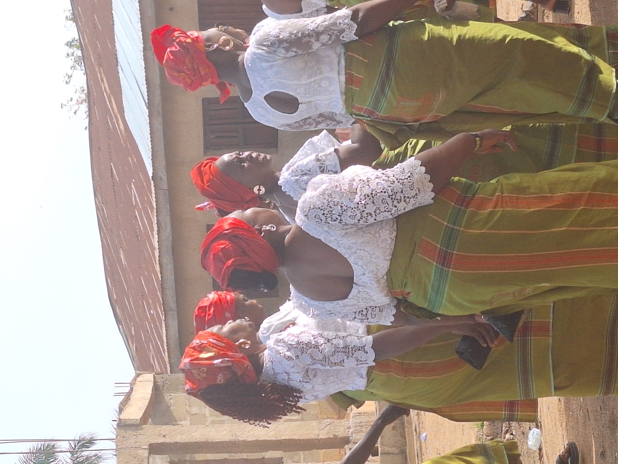 The image features at least five women dressed in traditional attire, captured outdoors in what appears to be a sunny daytime setting in Sabon Tasha, Nigeria. The women are arranged in a line or group, generally looking forward or slightly upwards, suggesting they are attending an event or posing.
Each woman is adorned in a white, lacy short-sleeved blouse paired with a long, olive-green wrapper (iro) featuring horizontal stripes in red, orange, and thinner lines of green or blue. Their heads are elegantly covered with vibrant red headwraps (gele), some of which are embellished with gold patterns. Several women wear large, gold-colored hoop earrings. One woman in the foreground is seen holding a dark object, possibly a phone or a small bag, in her hand.
The background reveals a simple building with light-colored walls, possibly concrete or mud brick, and a corrugated metal roof. Wooden window shutters are visible on the building's facade. The ground appears unpaved, consistent with a rural or semi-urban environment, with some sparse vegetation, including tree branches and palm fronds, visible against a bright, clear sky. The bright sky indicates it is clear and sunny during the day. The scene evokes a sense of community gathering or celebration.
There is no visible text in the image.