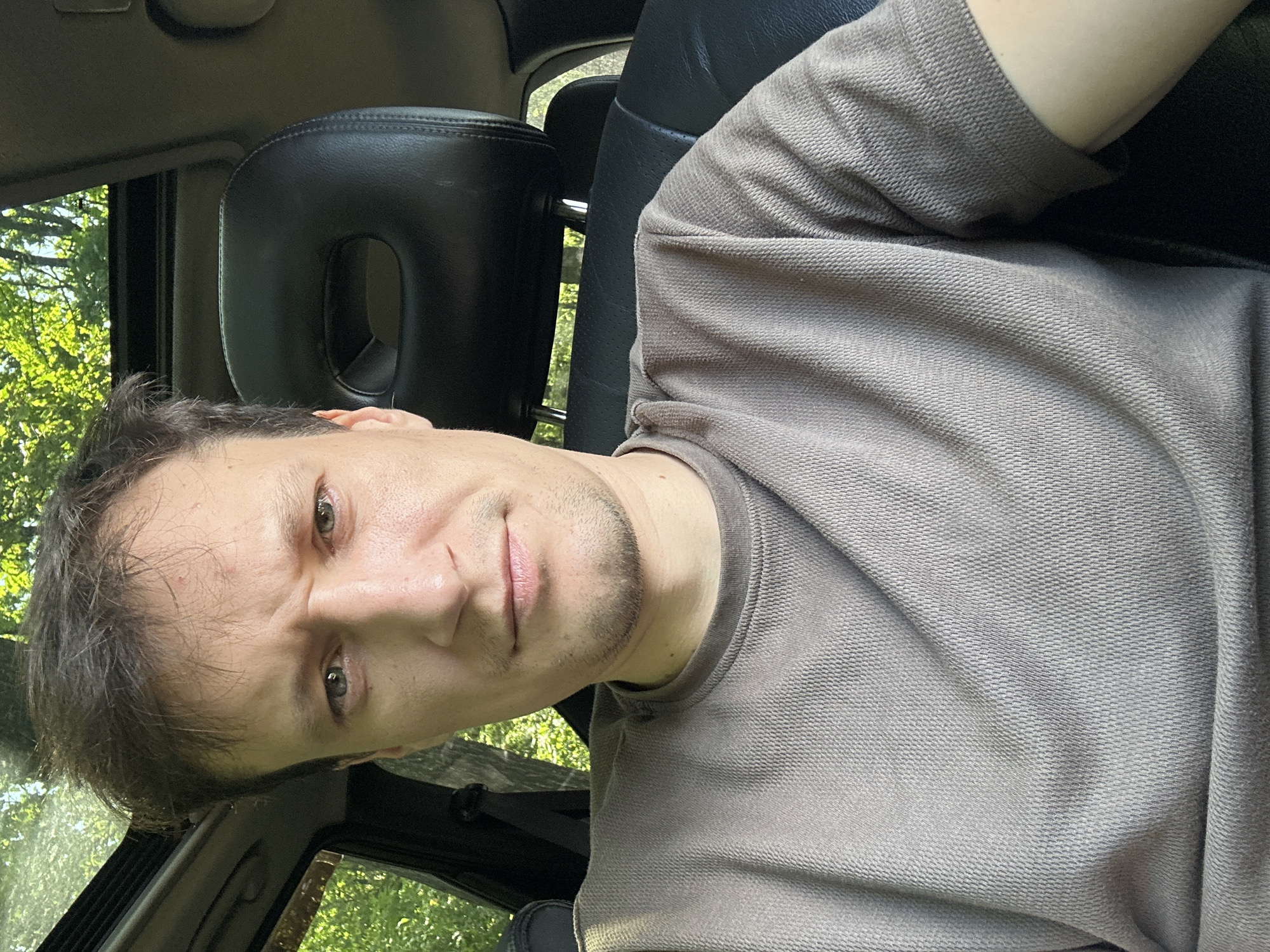 A man is seen from the chest up, sitting in the driver's seat of a car, with a sunroof open above him. He has short, dark brown hair, fair skin, and is looking directly at the camera with a concerned expression. He is wearing a brown, textured t-shirt. The interior of the car is visible, with black leather seats and headrests. The background shows a blurry view of green trees and foliage, suggesting the car is in a natural or wooded area. There is no indication of the specific location being Makiv, Ukraine.