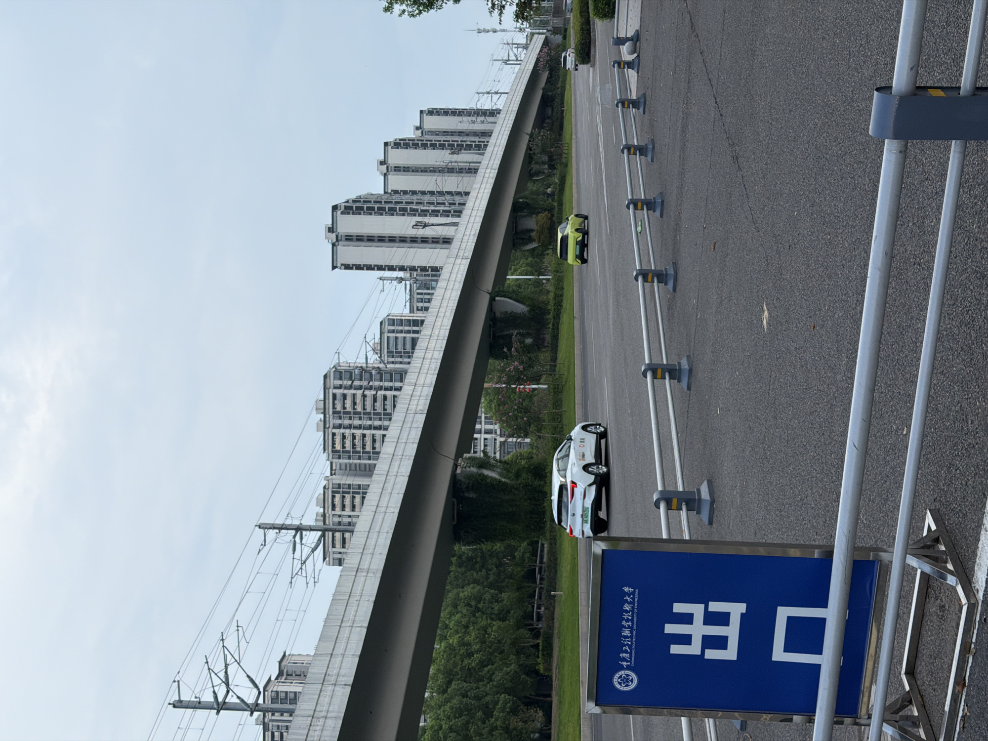 The image depicts an outdoor scene in Jiangjin, China, likely on a cloudy day, characterized by an elevated overpass with overhead power lines, and several residential buildings in the background. In the foreground, a blue sign with white Chinese characters "出口" (exit) and an emblem of "Chongqing University of Technology and Engineering" is visible. The sign is on a metal stand with a silver railing in front of it. The road surface is asphalt, with a white line marking a lane. Two cars are visible on the road; a white car to the left and a light green car further to the right. The environment appears to be urban or suburban, with lush green trees lining the side of the road and separating it from the overpass.