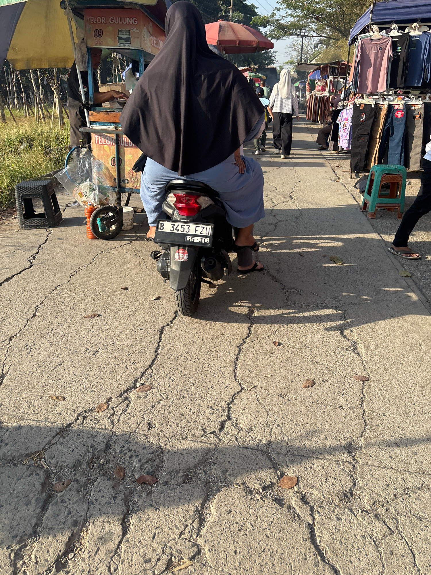 The image depicts a street scene in Telagamurni, Indonesia, with a focus on a person seated on a motorcycle. The individual is wearing a black hijab that covers their head and body, and light blue pants. They are positioned on a dark-colored motorcycle with a license plate displaying "B 3453 FZU" and the date "05-30". The motorcycle is parked on a cracked asphalt road.
In the background, several market stalls are visible. To the left, a food stall with a yellow awning sells "TELOR GULUNG" (rolled eggs). A vendor is partially visible behind the counter. Further down the street, another stall displays clothing on racks, with various t-shirts and pants available. Several people are walking or standing near the stalls. The environment appears to be a roadside market with a mix of vendors and pedestrians. The lighting suggests it is daytime, with shadows cast on the road.