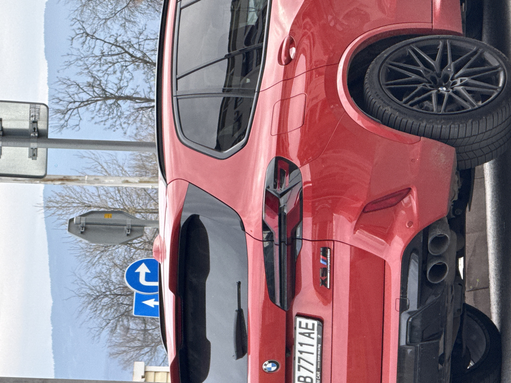 A red BMW X5 M Competition is parked near a street sign in Plovdiv, Bulgaria. The car has a black rear diffuser with quad exhaust pipes and a Bulgarian license plate reading "B 7711 AE". The location includes a street sign, bare trees, and a cloudy sky in the background. The car is parked next to a curb.