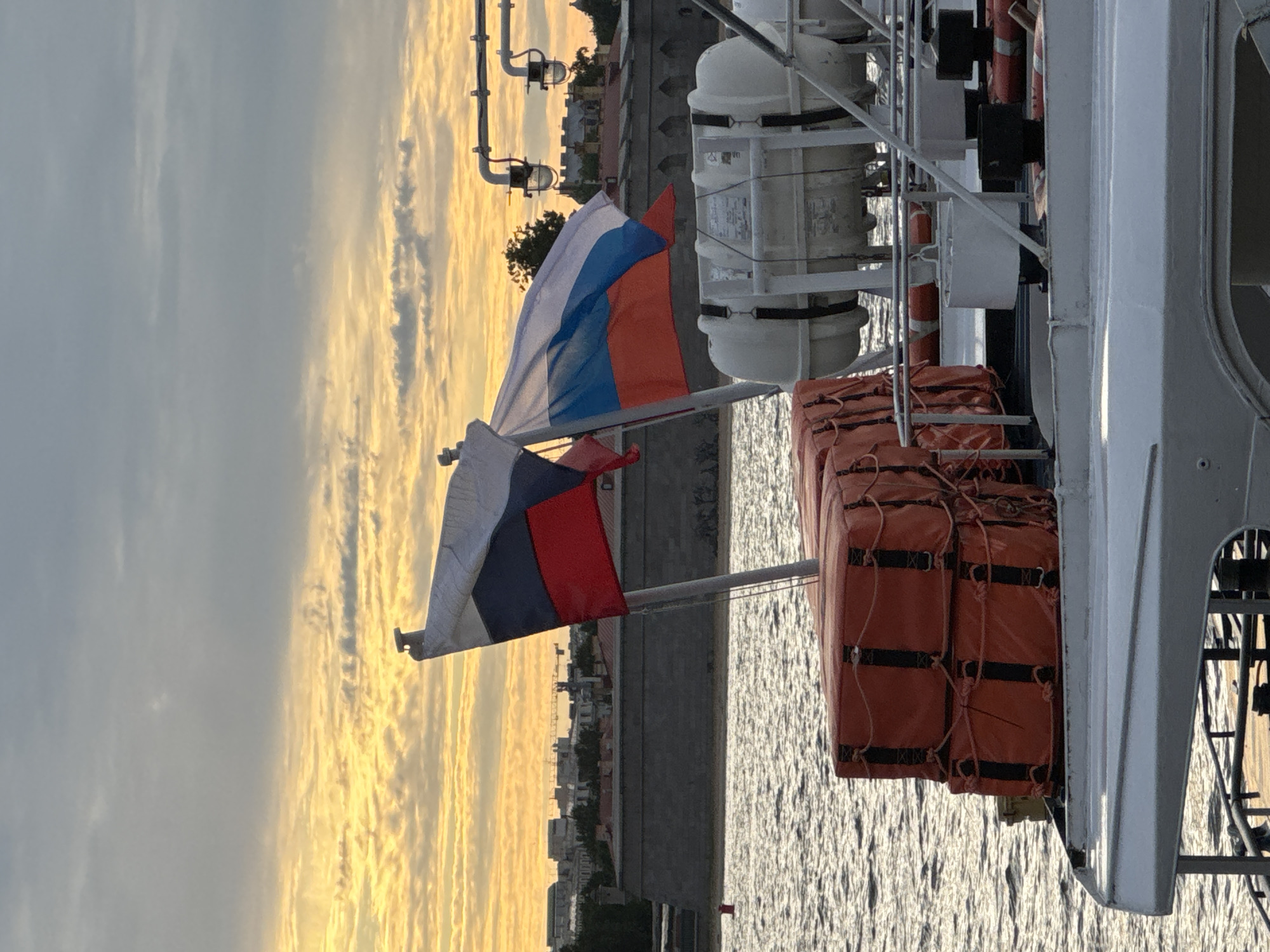 The image depicts a close-up view of the deck of a vessel on the Neva River in Saint Petersburg, Russia, at sunset. Two Russian flags are prominently displayed, fluttering in the breeze. In the foreground, several bright orange life rafts are stacked high, secured with ropes. Behind the flags, a white life raft canister is mounted on the deck. The background reveals a historic stone embankment, likely part of the Peter and Paul Fortress, with buildings and cranes visible in the distance. The sky is filled with a dramatic sunset, characterized by a vibrant orange and yellow glow illuminating the scattered clouds. The water of the Neva River reflects the colors of the sky. There are no people visible in the image.