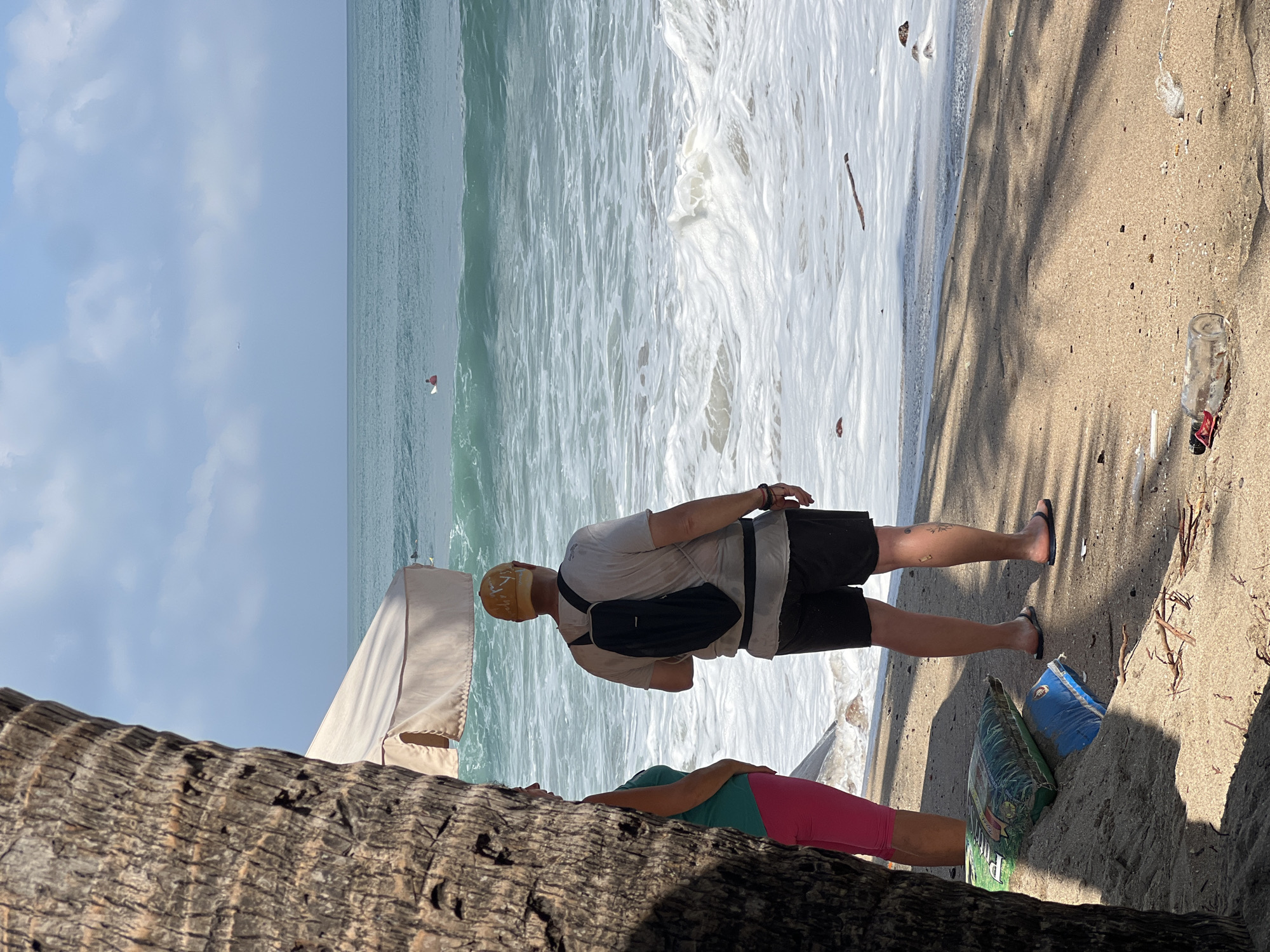 The image depicts a beach scene in Kuta, Indonesia, with two individuals standing by the ocean. A person wearing a yellow cap, a light-colored t-shirt, and black shorts is facing away from the camera, with a black backpack slung over their shoulders. To their left, partially obscured by a palm tree trunk, another person is visible from the waist down, wearing a green tank top and bright pink pants.
The ocean is a prominent feature, with waves breaking and white foam washing ashore. In the distance, a lone surfer can be seen on a wave. The beach itself is sandy, with some debris scattered on the ground, including plastic bottles and dried twigs. Two large bags, one green and one blue, are placed on the sand near the people. The sky above is partly cloudy. A beach umbrella, mostly out of frame, is visible behind the individuals. The overall impression is of a relaxed, outdoor setting on a sunny day at the coast.
