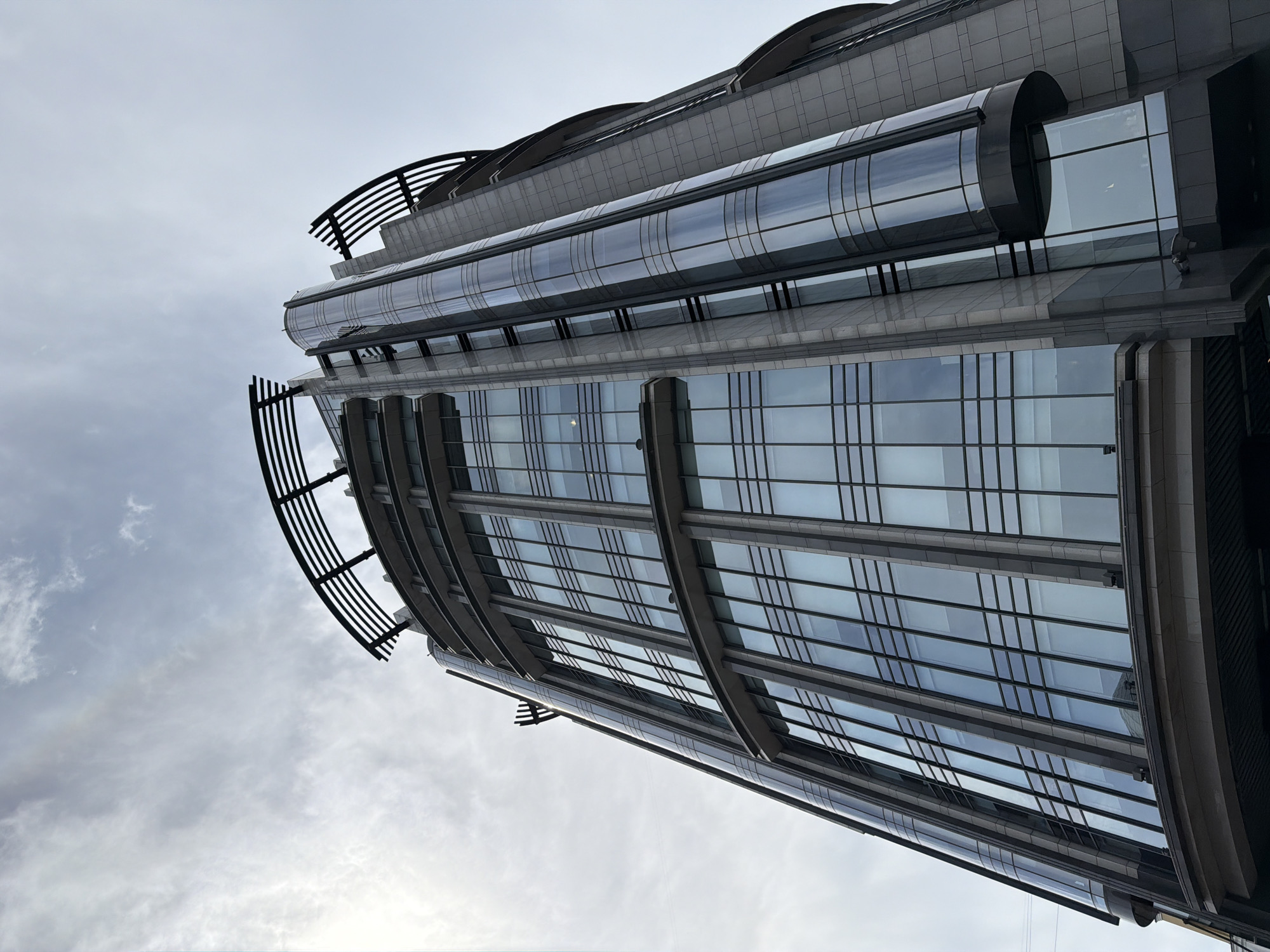 A low-angle, wide shot captures a modern, tall glass and stone skyscraper against a cloudy, overcast sky. The building, located in Moscow, Russia, features a curved facade with large, rectangular windows separated by dark mullions. A prominent, cylindrical glass section runs vertically along one side of the building, accentuating its modern design. The upper section of the skyscraper is adorned with curved, black metal structures that resemble arches. The lower portion of the building is darker, with a visible overhang. The sky is a pale gray, with scattered wisps of white clouds. The image is devoid of any people or vehicles.