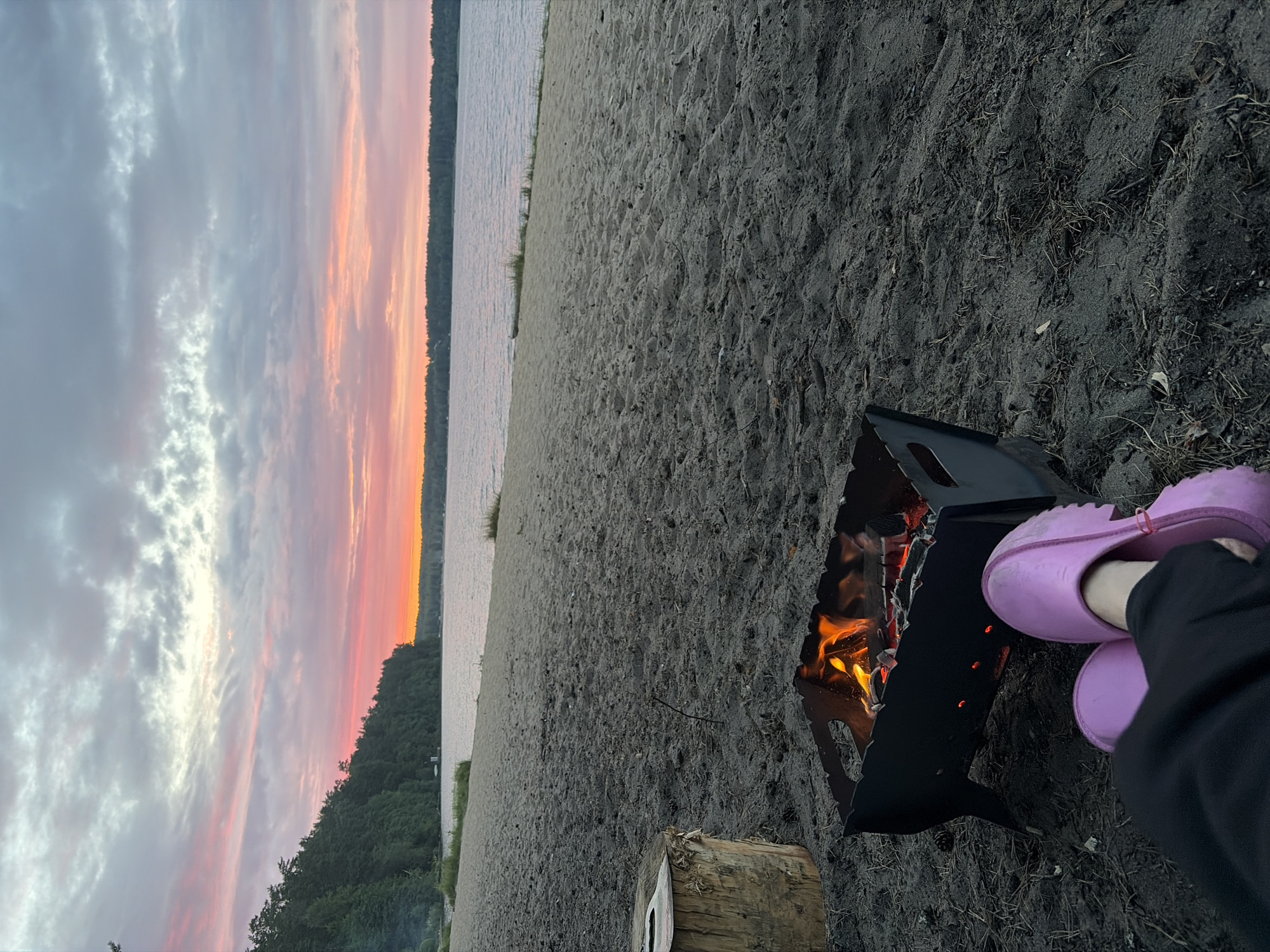 In Vaskelovo, Russia, a serene beach scene unfolds as dusk settles. A portable barbecue pit crackles with a vibrant fire, its flames casting a warm glow on the dark sand. Nearby, a tree stump serves as a rustic seat. In the foreground, a pair of pink clogs rests on the sand, hinting at the presence of someone enjoying the peaceful evening. The tranquil lake stretches out to the horizon, framed by a dense forest of green trees along the left bank. The sky above is a canvas of soft, pastel colors, with hues of pink and orange blending into the gray clouds, a hallmark of a Vaskelovo sunset.