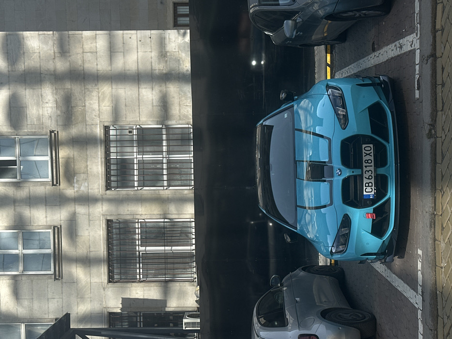 A turquoise (or light blue) BMW G-series M3/M4, bearing Bulgarian license plate "CB 6318 XO", is parked in an outdoor paved parking space in Sofia, Bulgaria. The vehicle features a black roof, two black stripes centrally located on its hood, and a visible red tow hook attachment on the lower left of its front bumper. It is positioned facing the camera. To its immediate left, a silver Fiat 500-type vehicle is partially visible, parked in an adjacent space. To its right, a dark-colored sedan is partially visible. The parking surface is composed of light-toned pavers with white painted demarcation lines. A raised curb separates the parking area from a foreground paved section.
Behind the parked cars, a dark, reflective barrier or wall extends horizontally. Above this barrier, a multi-story building with a light gray, possibly stone or concrete, facade is visible. The building features several rectangular windows with white frames; the lower-level windows are fitted with dark metal security grilles. An external air conditioning unit is mounted on the building's facade on the far left. Distinct, tree-like shadows are cast across the building's surface, indicating direct sunlight from an unseen source. The scene is illuminated by natural daylight.