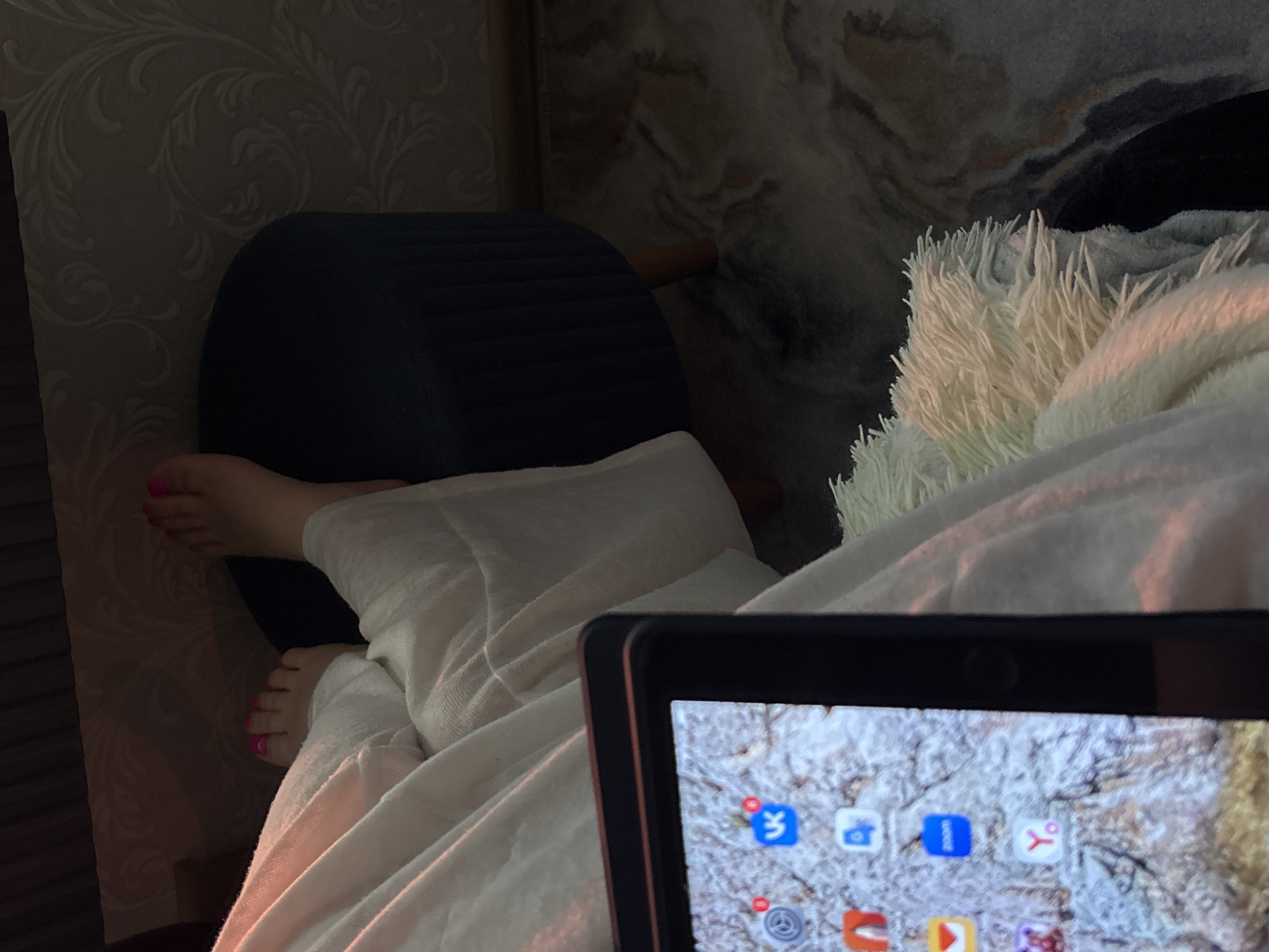 The image shows a person relaxing indoors. The person's feet, with pink nail polish, are resting on a dark blue, ribbed ottoman or stool. The person is covered in a white blanket and using a tablet. A fluffy white pillow is visible next to the blanket. The background includes wallpaper with a textured pattern and what appears to be a patterned wall covering or headboard.
It is impossible to confirm the location in Chaikovsky, Russia without additional information or context in the image.