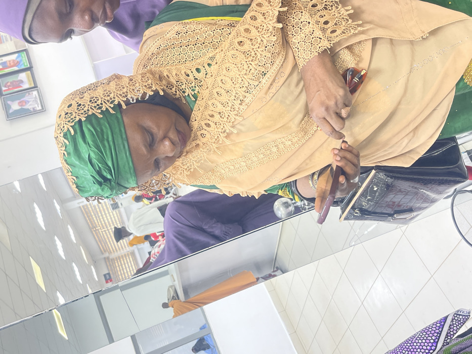 An indoor scene in Zaria, Nigeria, features a woman, likely middle-aged, positioned centrally on the right, looking downwards at a dark-colored smartphone held in both hands. She wears a green headwrap and a beige lace-edged veil over a green garment. Her left wrist displays multiple silver-colored bangles. A pair of brown-framed eyeglasses is held in her right hand near the phone. A dark brown, crocodile-patterned handbag with a gold-colored clasp is visible below her hands. The upper right corner shows the partial view of another woman, wearing a purple head covering, observing the main subject. The left side of the image is dominated by a large mirror, reflecting an interior space with white walls, a light-colored tiled floor, and several individuals, including one in an orange garment and others further back. On the wall in the upper right background, three framed portraits of men in traditional attire are mounted. The environment is brightly illuminated by overhead artificial lighting.