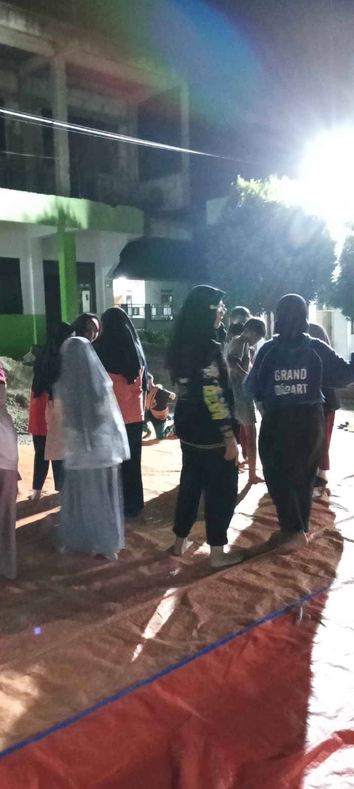 A group of people, mostly women wearing hijabs, stand barefoot on a shiny, reddish-brown surface. The setting appears to be outdoors at night, illuminated by bright overhead lights. In the background, a building with green accents is visible. Some individuals are facing away, with "GRAND DEPART" printed on the back of one's clothing. The scene has a casual, somewhat blurry quality due to the low lighting.