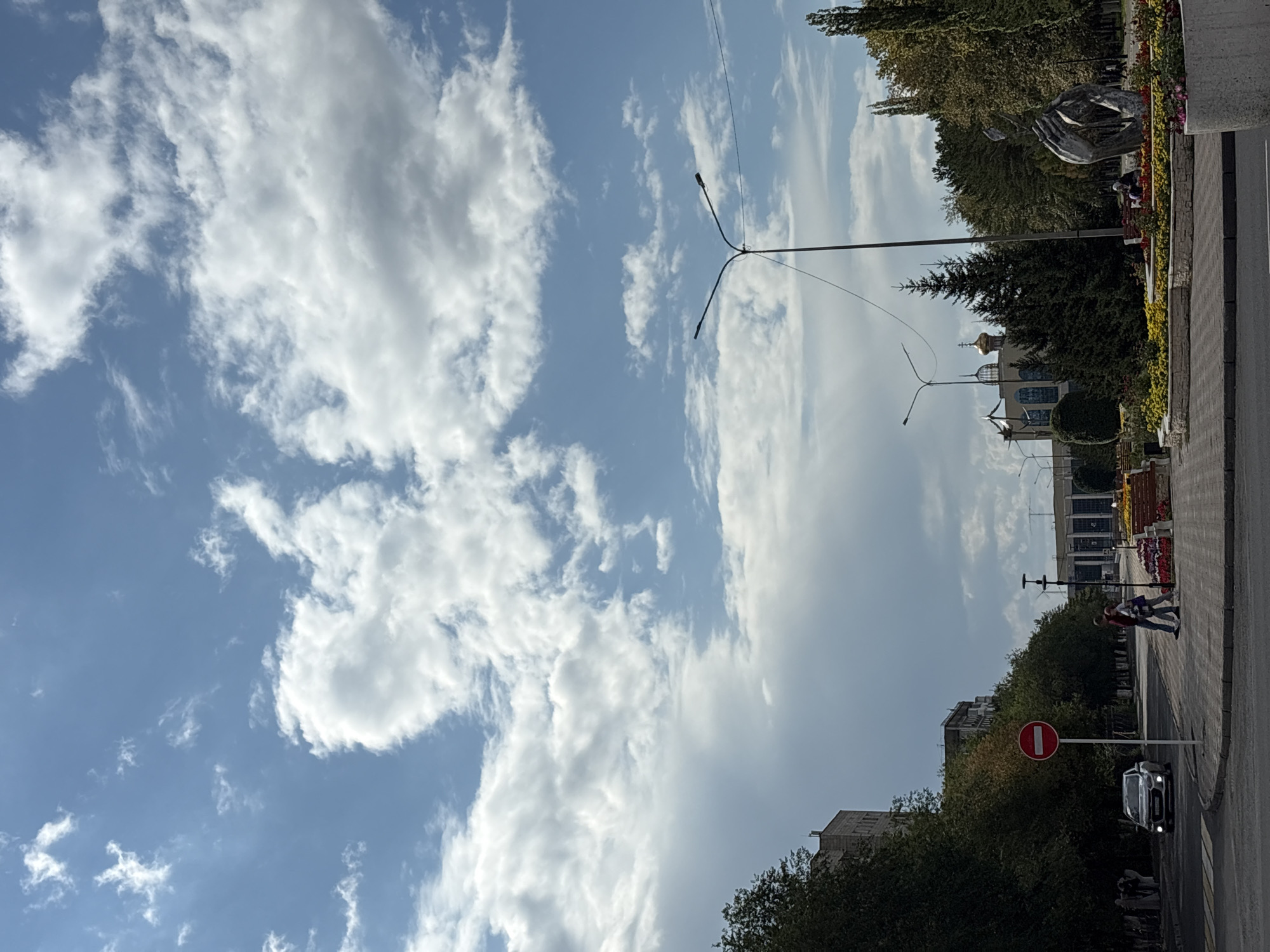 This is a street-level, eye-level photograph of a city scene in Pavlodar, Kazakhstan, on a day with a partly cloudy sky. The view is looking down a wide street lined with trees.
In the foreground, a grey sedan is driving on the left side of the road, and two people are walking on the sidewalk. A red and white "no entry" sign is prominently placed on the sidewalk. A sculpted monument of two hands embracing is visible in the middle ground, surrounded by flower beds. Behind the monument, there are several trees and a building with a golden dome, likely a religious structure. A lamppost with two lights stands tall in the middle of the frame. Further down the street, a large building with a modern facade is visible. The sky is filled with a mix of white and grey clouds.