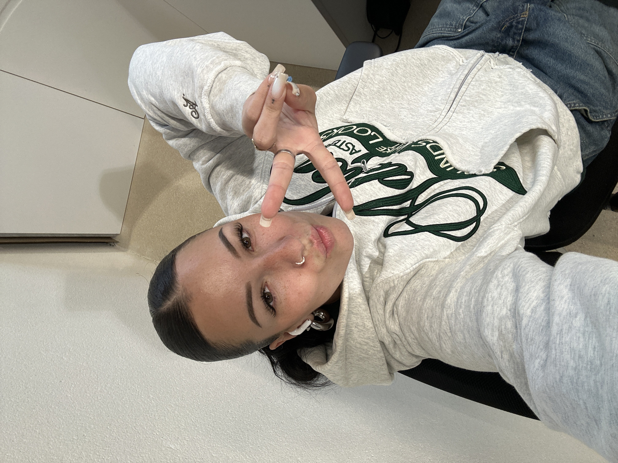 A woman is posing for a selfie. She is wearing a gray hooded sweatshirt and blue jeans. She is holding up two fingers in a peace sign. She has long black hair pulled back from her face and is wearing earrings. She has a nose ring and long fingernails. The background is a plain white wall and a black chair. There is no evidence in this image to indicate the location is in the city of Paris, France.
