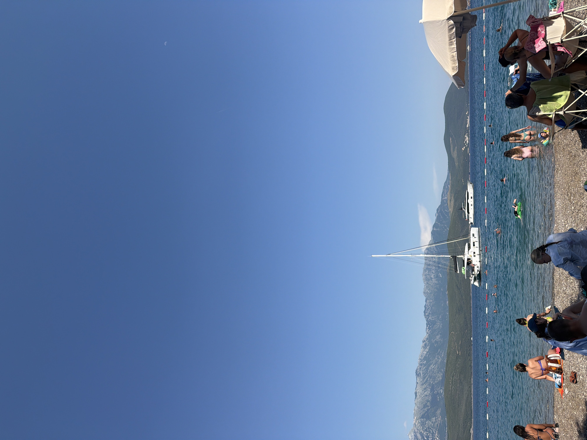 In Radovići, Montenegro, a serene beach scene unfolds under a clear blue sky. The foreground is occupied by a diverse group of people enjoying the pebbled shoreline and the tranquil sea. Some are seated in beach chairs, others are sunbathing on towels, and a few are wading into the cool, clear water. The middle ground features several individuals swimming and playing in the sea, with a bright green inflatable toy visible. Further out, a majestic white catamaran with multiple sails is anchored, and a sleek white motor yacht is also present. The background is dominated by lush green hills that rise to meet imposing, rugged mountains under a vast expanse of sky. The image captures a leisurely summer day at the coast, with the natural beauty of Montenegro providing a stunning backdrop.