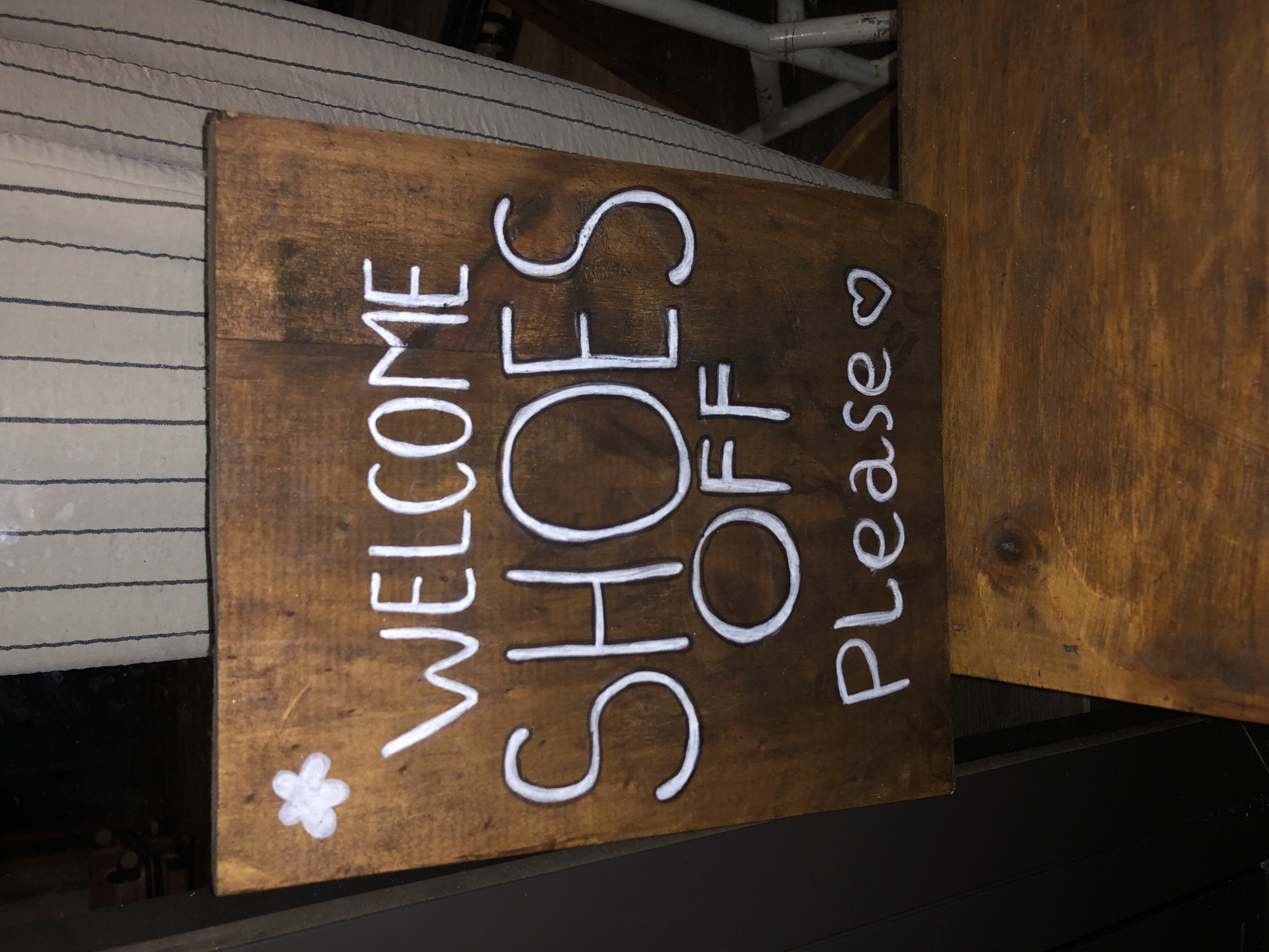 The image shows a wooden sign with white lettering that reads "WELCOME SHOES OFF Please". There is also a white flower drawing at the bottom left of the sign and a heart drawing at the top right. The sign is positioned vertically on a wooden surface, likely as a door sign. The background is blurred, but appears to be an interior setting with some light fixtures and a striped fabric. The overall mood of the image is welcoming and informative. The sign's message suggests a request for visitors to remove their shoes before entering, indicating a culturally sensitive environment or a preference for a clean interior.