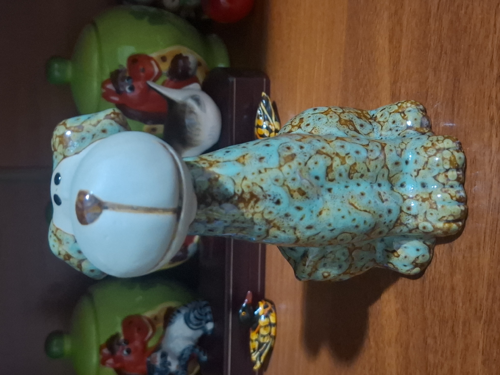 A ceramic figurine of a dog with a light blue and brown speckled glaze sits on a wooden surface. The dog figurine is in the foreground, facing forward. Behind the dog, on a shelf, are several other decorative items. To the left, a green ceramic jar with a lid is visible, with a small figurine of a red horse and a gray and white cat in front of it. To the right of the dog, another green ceramic jar with a lid is partially visible. Further to the right, a red sphere, possibly a ball or another decorative item, is visible. A small decorative duck figurine with yellow and black markings and a red beak is placed on the wooden surface to the left of the dog figurine. The lighting suggests an indoor setting, possibly a home or a shop. There are no people or animals present in the image. The scene is static and appears to be a display of decorative objects. The location is not specified.