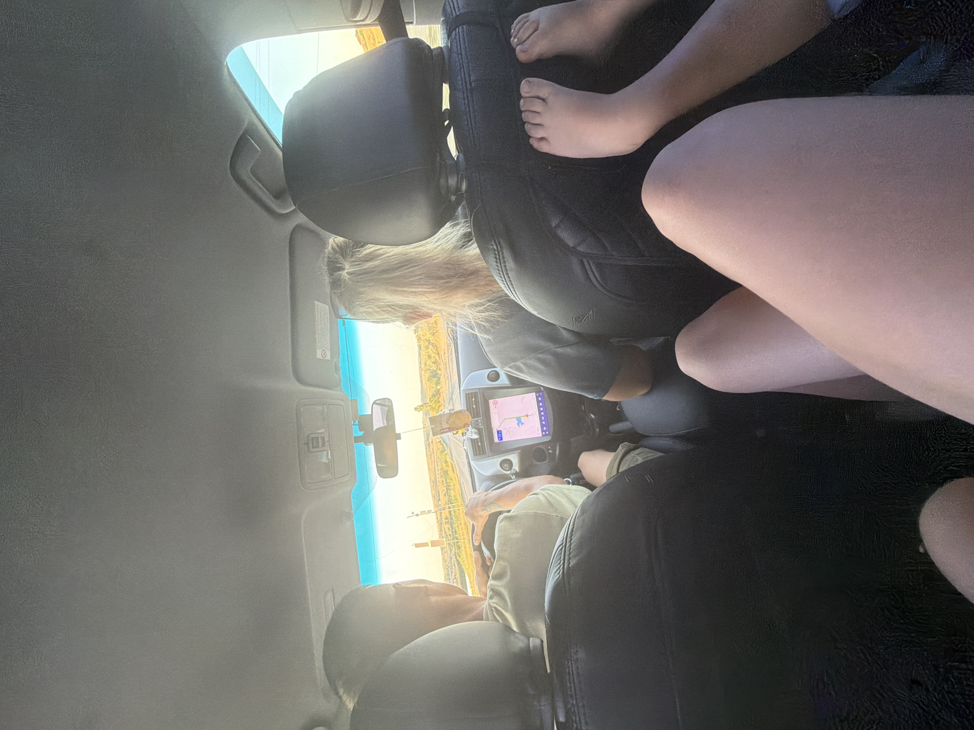 This is a low-angle, interior shot taken from the back seat of a car, looking forward. The image captures a family on a road trip. In the driver's seat is a man with short hair, wearing a light green t-shirt, his hands on the steering wheel. Beside him, in the passenger seat, is a woman with blonde hair tied in a ponytail, wearing a grey t-shirt. The car's navigation screen is visible on the dashboard, displaying a map. In the foreground, the legs of at least two people are visible, one set of small feet resting on the back of the front passenger seat. The sunlight streams in through the windshield and side windows, indicating it is daytime. The background outside the car shows a rural landscape with fields and a road stretching into the distance. The scene appears to be in the city of Belogorki, Russia.
