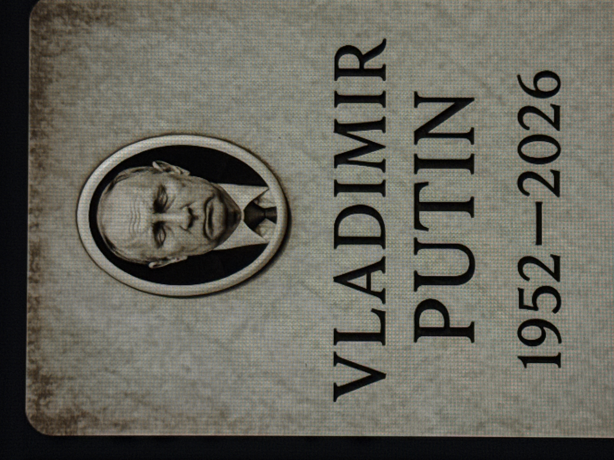 The image displays a tombstone-like monument with a portrait of Vladimir Putin in an oval frame at the top. Below the portrait, the text "VLADIMIR PUTIN" is inscribed, followed by the dates "1952-2026". The monument has a weathered, stone-like texture. This monument is located in the city of Bratislava, Slovakia, specifically at Hodžovo námestie.
