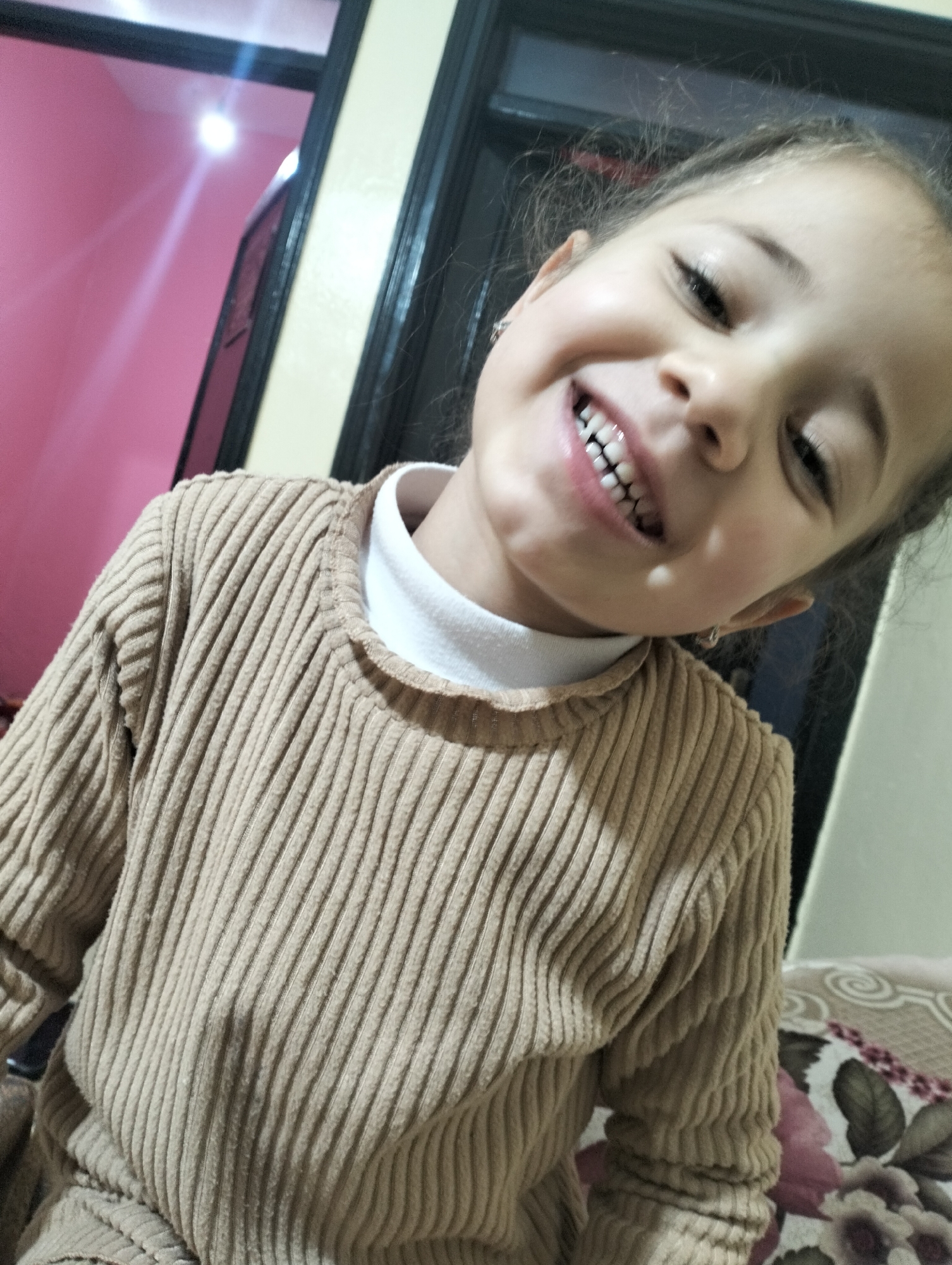 A young child with brown hair, a tan complexion, and visible upper teeth smiles at the camera. The child wears a beige corduroy-like shirt over a white undershirt. The background includes a pink wall with a door frame. The bottom of the frame shows part of a bed with a floral pattern. The photo appears to be taken indoors. There is no information in the image suggesting its location in Ouaouizert, Morocco, and no additional details can be determined from this picture.