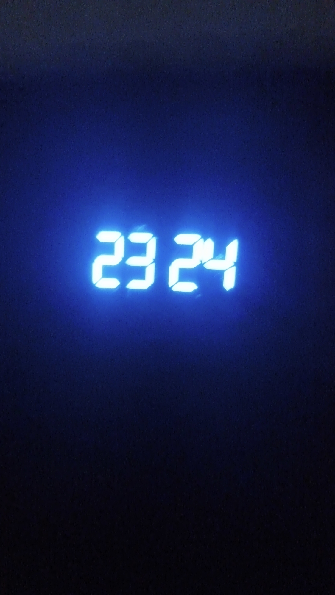 The image displays a digital clock showing the time "23 24" in bright, glowing blue numbers. The numbers are rendered in a segmented style, characteristic of digital displays. The background is extremely dark, appearing as a deep blue or black, suggesting a low-light environment, likely nighttime. The blue light from the digital display subtly illuminates the immediate surroundings, creating a faint gradient from dark blue to slightly brighter blue around the numbers. There are no other objects, people, or discernible activities present in the frame. The overall impression is minimalist and focused solely on the time display in a dark setting. The image itself appears to have some digital noise or graininess.