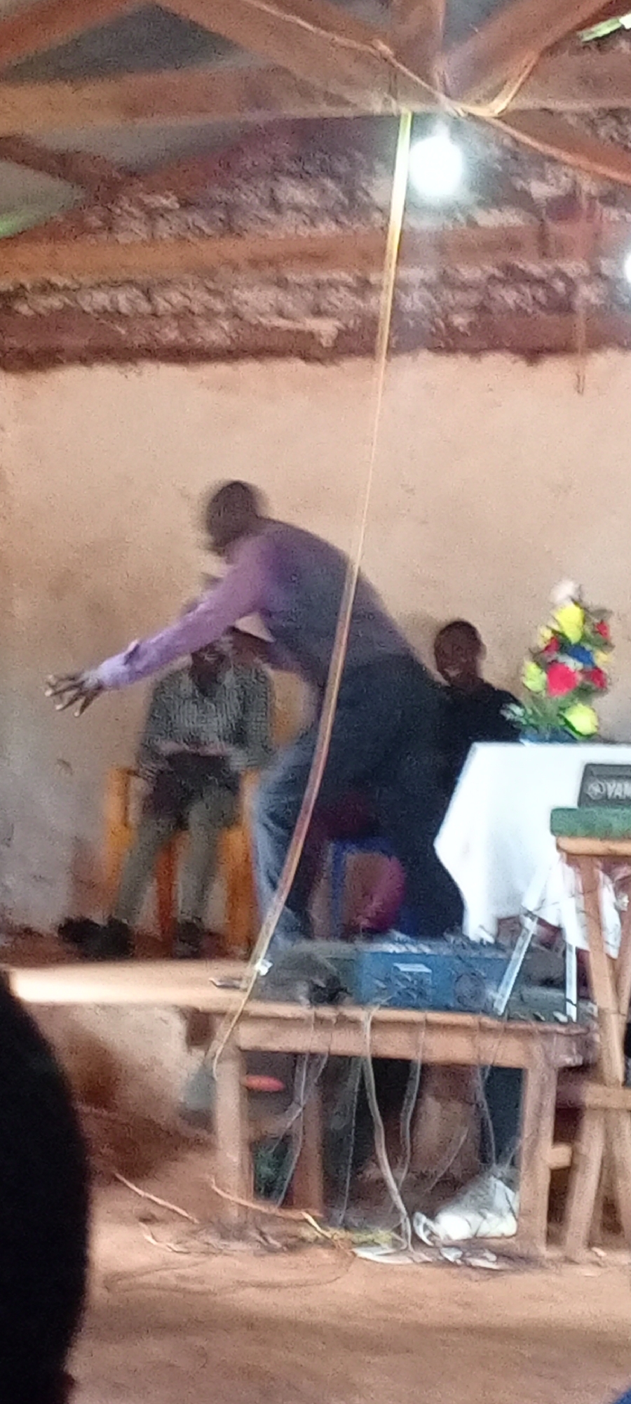 The image depicts a scene inside a building in Ngalula, Democratic Republic of the Congo. A man in a purple shirt and dark pants is gesturing with his left arm extended. He is standing on a raised platform made of wooden tables. A soundboard and various electrical cords are visible on and under the table. To his left, a man is seated on a yellow chair. Behind the man on the platform is another man dressed in black, sitting behind a table with a white tablecloth and a vase of colorful artificial flowers. A light bulb hangs from the ceiling, which is constructed of wooden beams and a textured, light-colored material.