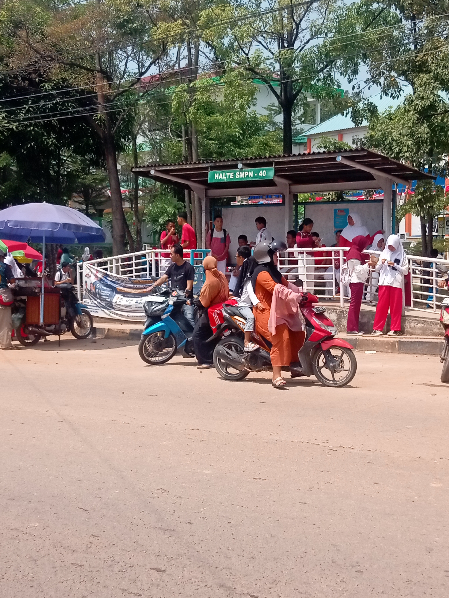 The image captures a street scene in Batam, Indonesia, likely near a school, indicated by the "HALTE SMPN - 40" sign on the bus stop. Several people are gathered around a bus stop shelter, with some waiting and others engaged in conversation. Two motorbikes are prominently featured in the foreground. One blue motorbike is ridden by a man in a black t-shirt, with a woman wearing an orange hijab as a passenger. Another red motorbike has a woman in an orange hijab and a child on it, with the woman appearing to be wearing a pink headscarf. A food cart with a blue umbrella is visible on the left side of the image, suggesting a commercial area. Trees and buildings form the backdrop, creating a typical urban environment in a tropical setting. The overall atmosphere appears to be one of everyday activity, with people commuting and interacting.