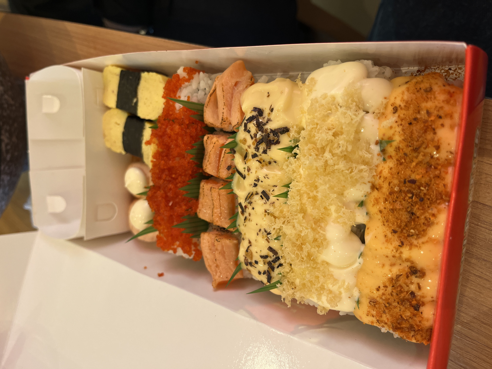 A box of sushi rests on a wooden surface. The sushi is arranged in four distinct varieties from left to right. The first type features egg and seaweed. Next, there is sushi covered in orange roe, interspersed with small white spheres. The third sushi type is topped with salmon slices, green leaves, and small dark sprinkles. The final variety has a creamy sauce and a sprinkle of brown crumbs. The box is white with a red edge, and the wooden surface appears to be a table. The specific location in Lhokseumawe, Indonesia cannot be determined from the given information.