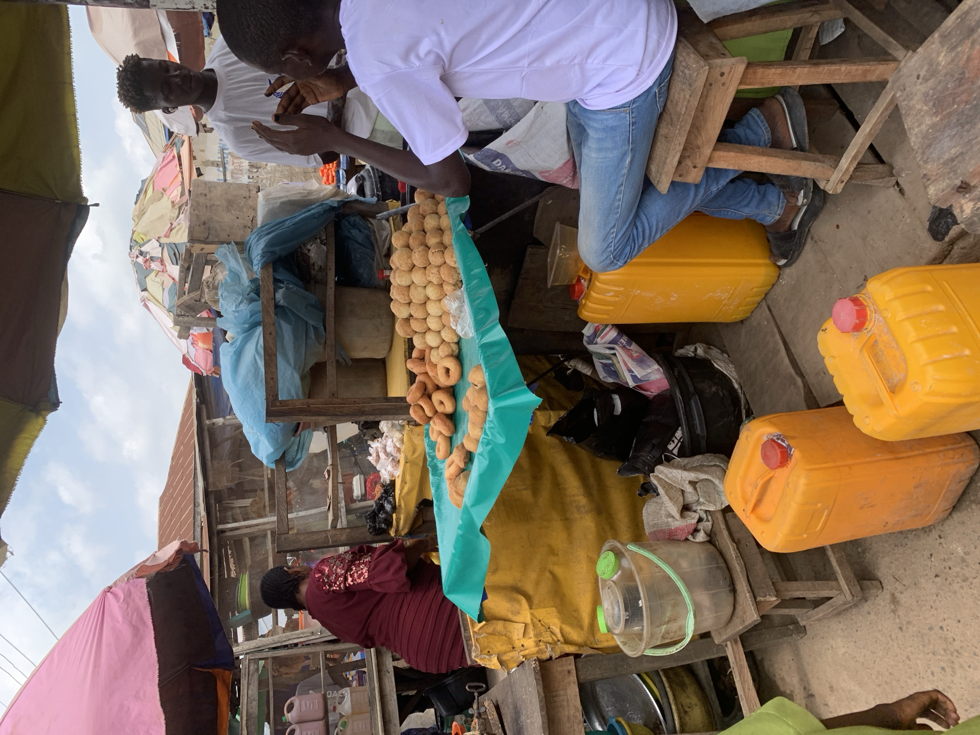 The image depicts a bustling street scene in Shagamu, Nigeria. Two young men in white shirts are seated on wooden structures; one wears jeans and sits higher, while the other sits lower. A string of round, fried pastries hangs from a turquoise covering attached to a market stall below them. Next to the pastries, a woman in a maroon shirt stands behind the stall. Several yellow jerrycans are visible near the seated men. The scene is crowded with objects and stalls covered with various colored tarps.
