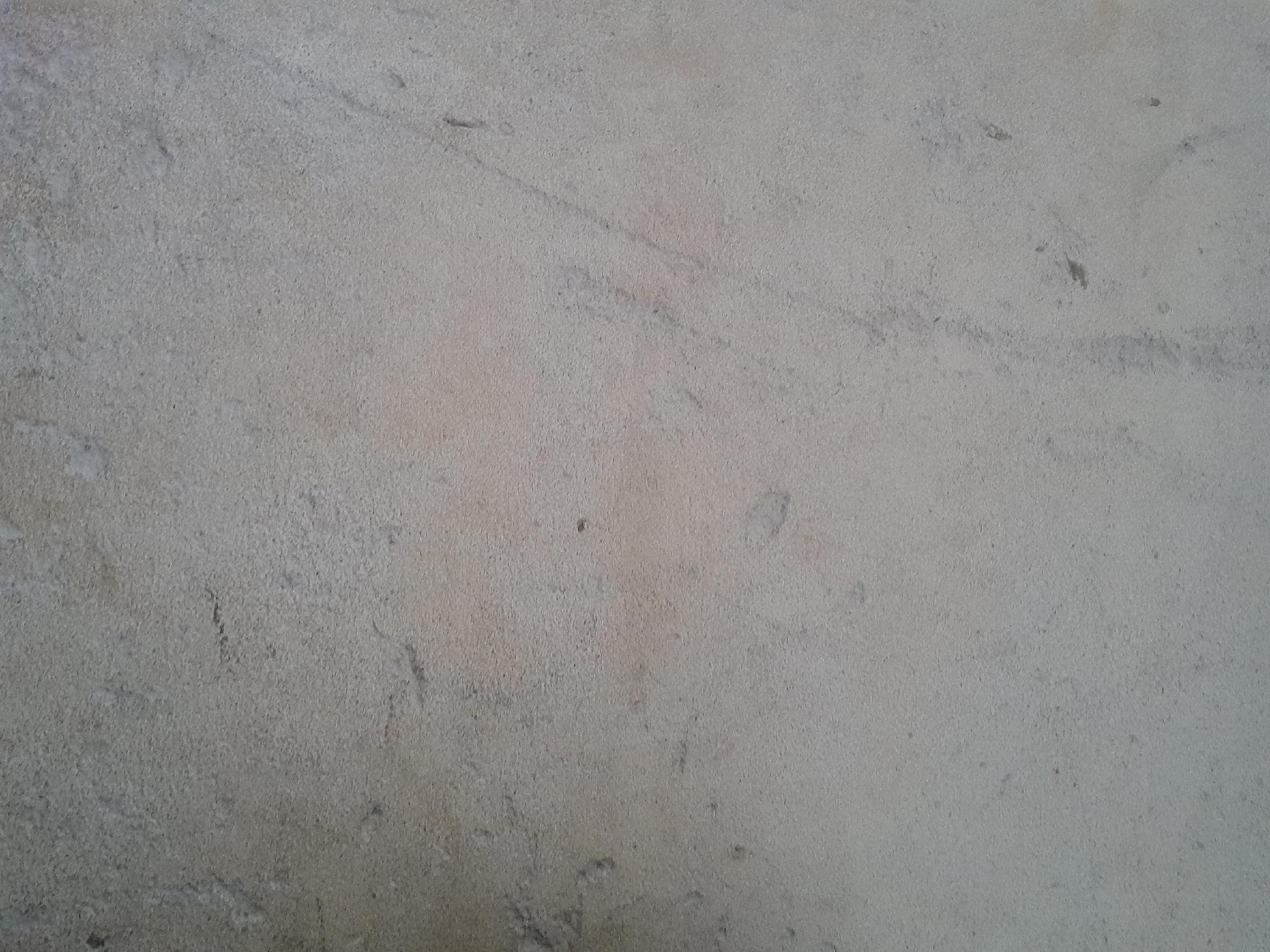 The image displays a close-up, static view of a textured surface. The surface is predominantly light grey with subtle beige and off-white undertones, exhibiting a fine-grained, irregular texture across the entire frame. Scattered across this surface are sparse, indistinct darker grey smudges, minor abrasions, and faint linear striations. There are no discernible people, specific objects, or environmental features visible that would allow for precise geographical identification within the city of Ifo, Nigeria. The scene shows no actions or interactions. The illumination appears even and diffuse.
