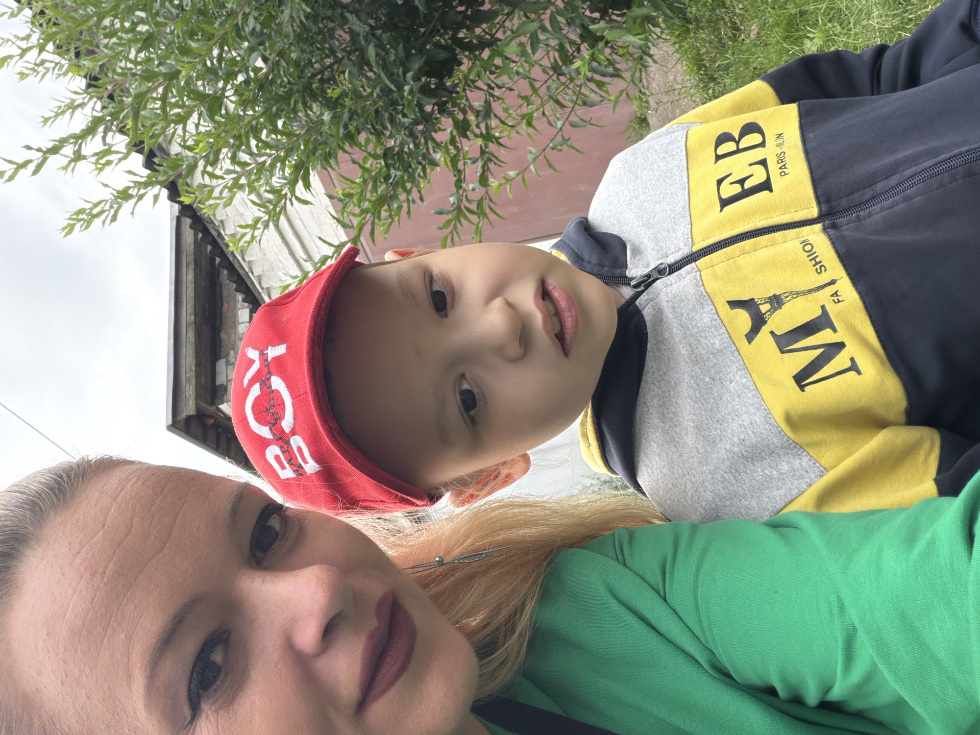 An outdoor, low-angle, front-facing selfie depicts a woman and a young boy. The woman, on the left, has blonde hair pulled back, is wearing a green top, and has makeup applied. Her face is close to the camera, showing her forehead, eye, and lips. The boy, positioned to her right, is wearing a red baseball cap with white lettering that appears to say "BOY". He is also wearing a zippered hoodie with yellow and gray panels, featuring black lettering that says "MY EB PARIS-LON". The background suggests an outdoor setting with a building and greenery, possibly in a residential area. The sky is overcast. The scene is likely in Chernihiv, Ukraine, given the context.