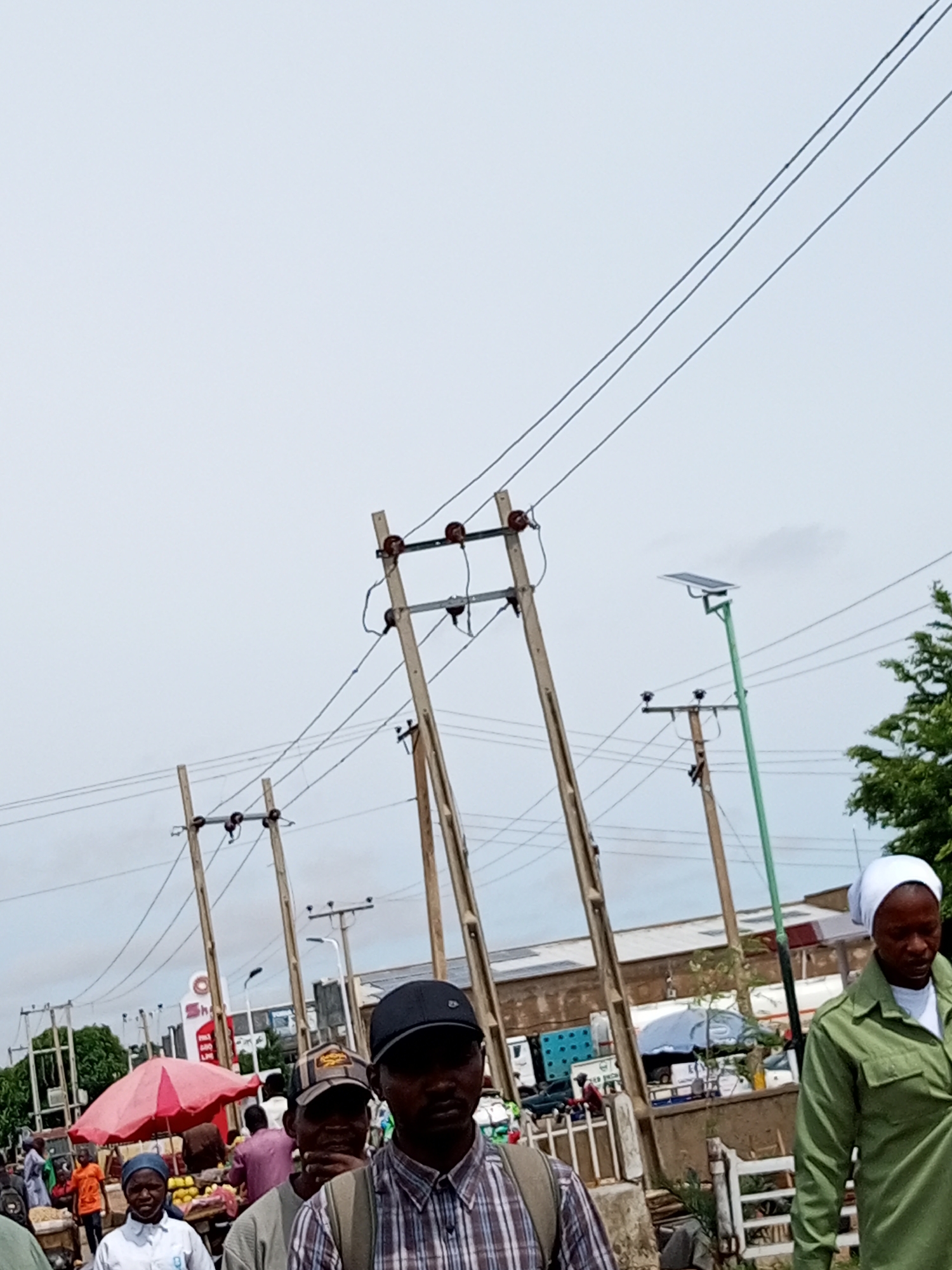 The image depicts a bustling outdoor market scene in Tumfure, Nigeria, under an overcast sky. In the foreground, a man wearing a plaid shirt and a dark baseball cap is visible, his face partially obscured. To his right, another man, partially out of frame, is also wearing a cap. Further into the scene, a person in a green uniform and a white head covering walks from right to left, looking down.

Behind them, a lively market unfolds with various stalls and vendors. A bright pink umbrella shades a section of the market, where fruits and other goods are displayed. Several people are seen browsing and interacting at the stalls. Power lines crisscross the sky, supported by wooden poles, and a solar-powered street light stands tall on a green pole. In the background, buildings and parked vehicles, including trucks, are visible, suggesting a commercial area. The overall impression is one of a dynamic and active marketplace.