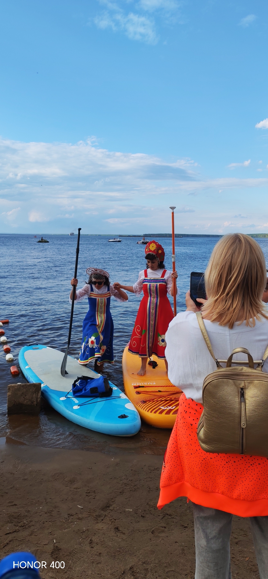 Two young women in traditional Russian costumes are standing on paddleboards in the Kama River near Chaikovsky, Russia. One woman is wearing a blue dress with floral embroidery and a white lacy overlay, and the other is wearing a red dress with decorative trim and a red headdress. Both women are holding paddles. In the foreground, a woman with blonde hair is photographing them with her phone. The background shows a blue sky with scattered clouds and a distant shoreline. The water is calm, and a few boats are visible on the river. The scene appears to be a recreational outing or a cultural event.