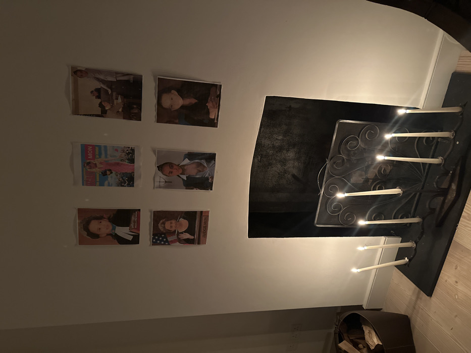 An interior room in London, United Kingdom, is depicted, illuminated primarily by six lit white taper candles positioned on and around a dark metal fireplace screen. The screen, featuring decorative scrollwork, rests on a dark hearth in front of a rectangular, dark-toned fireplace opening. Above the fireplace, six printed images or photographs are affixed to a light-colored wall, arranged in two horizontal rows of three. The top left image shows a child in a barrister's wig and gown. The top center image displays a woman in a pink dress, reminiscent of a magazine cover. The top right image features an adult and a child in what appears to be a professional setting. The bottom left image portrays a woman with an American flag background, labeled "JUDGE MON." The bottom center image shows a woman in an open blazer. The bottom right image shows a woman seated with her hands clasped. To the left of the fireplace, a brown, possibly leather, container holds several pieces of firewood on a light-colored wooden floor. The room's baseboards are dark-colored.