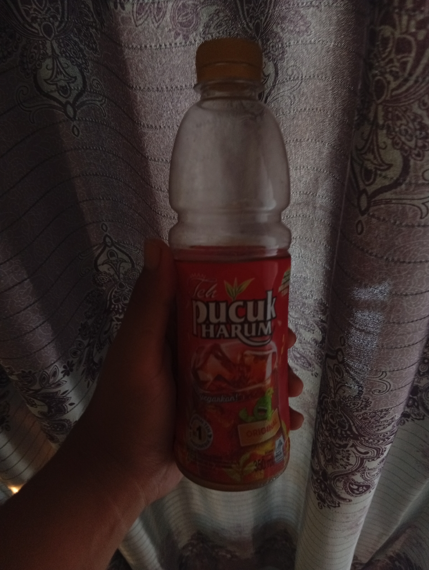 A hand holds a plastic bottle of Teh Pucuk Harum. The bottle has a yellow cap and a red label with green tea leaves. The background is a patterned curtain. The image is taken indoors.
