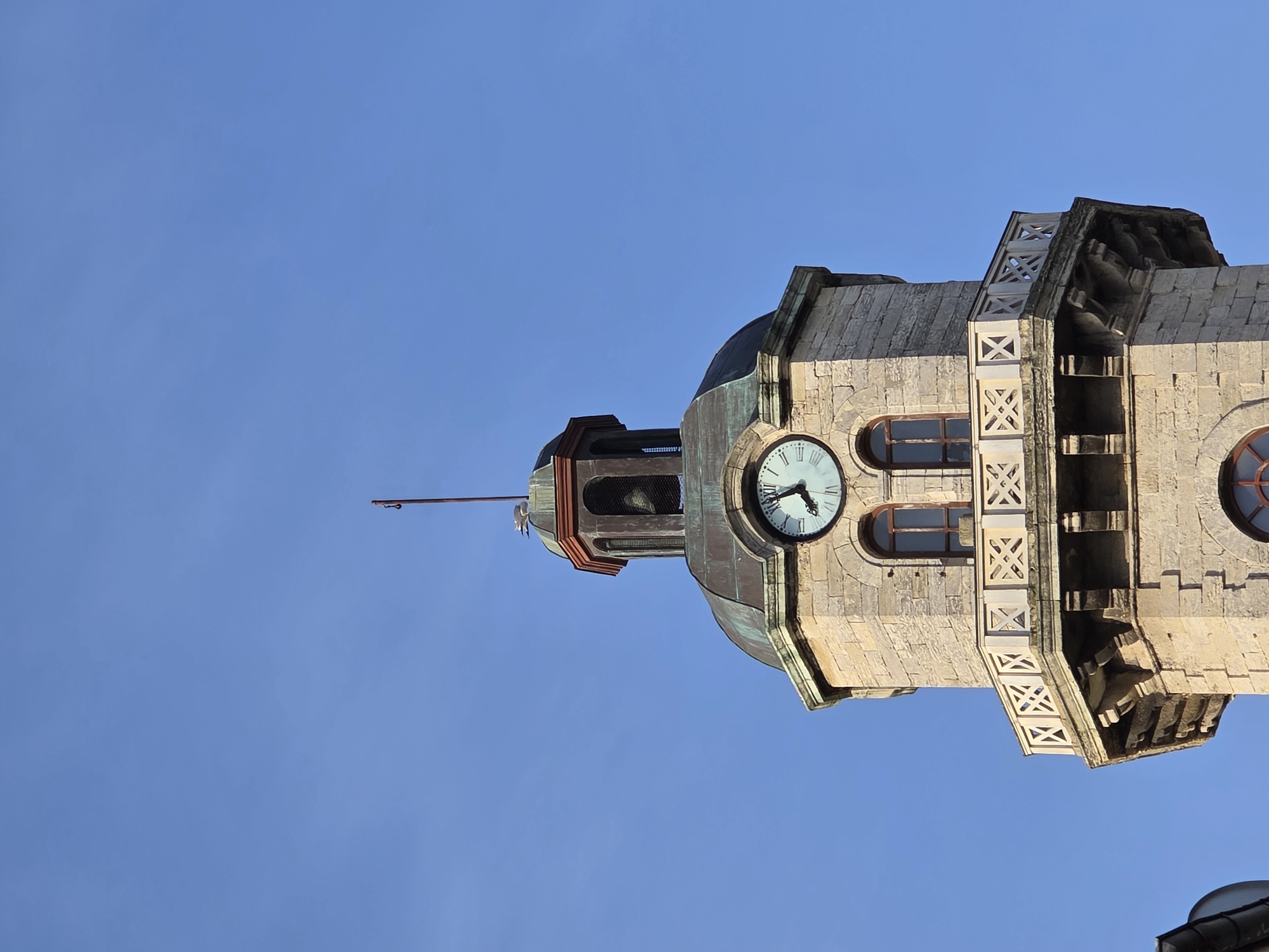 The image is a low-angle shot of the Saint Athanasius Church clock tower in Varna, Bulgaria, set against a clear blue sky. The tower is constructed of light-colored stone and features a clock face on one side, which shows the time at approximately 10:43. Windows with dark wooden frames are also visible on the tower. The tower is topped with a dome-shaped roof, a small open bell tower, and a spire. The tower's upper section is adorned with decorative stone lattice work. The lower right corner of the frame shows a portion of a dark roof, suggesting other buildings in the vicinity.