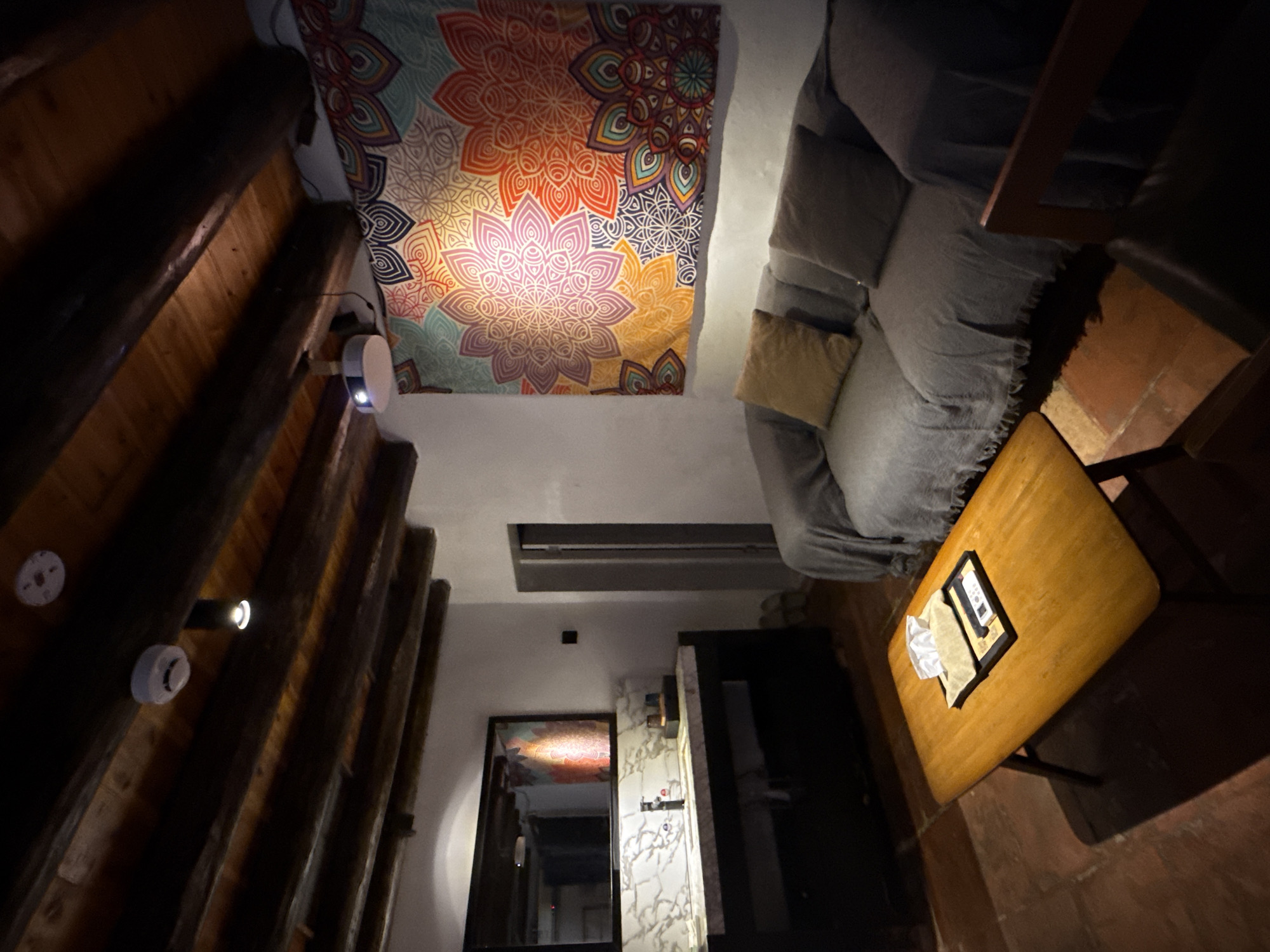 This image depicts a cozy living room or lounge area within a hotel room or private residence in Longgang District, China. The room features exposed wooden beams on the ceiling, adding a rustic touch. A projector is mounted on the ceiling, casting an image onto a vibrant, mandala-patterned tapestry hanging on the wall. In the foreground, a warm-toned wooden coffee table holds a tissue box and a remote control. A gray sofa, covered with a textured throw blanket and adorned with cushions, occupies the middle ground. To the left, a vanity area is visible, with a large mirror above a countertop that houses a sink, faucet, and various toiletries. The flooring appears to be terracotta tiles. The overall atmosphere is one of comfort and relaxation.