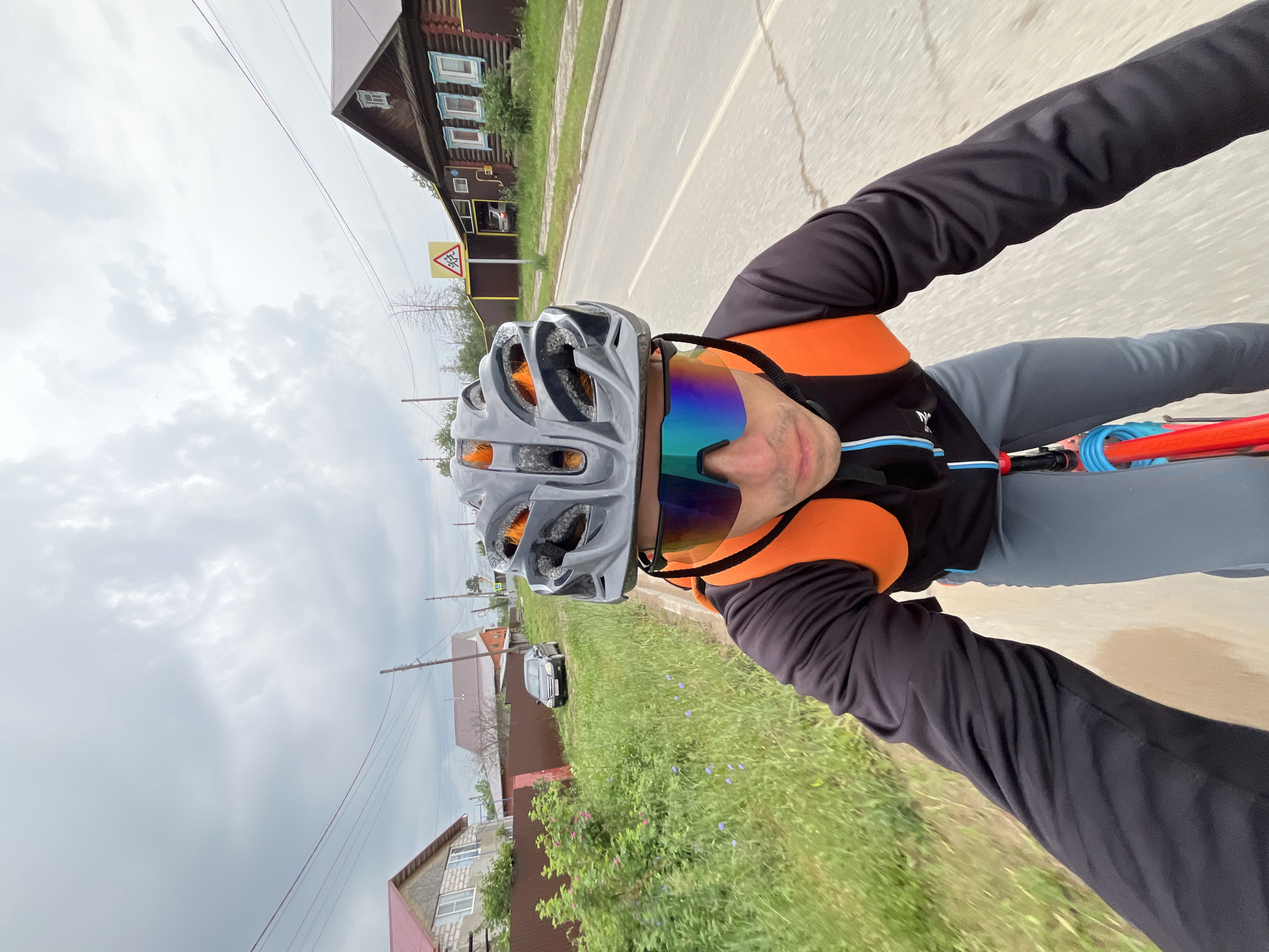 A man is riding a bicycle on a paved road in a rural area of Chaikovsky, Russia. He is wearing a black long-sleeved shirt, grey pants, an orange backpack, a grey helmet, and tinted cycling glasses. The road is lined with houses, fences, and green grass. There are power lines overhead. The sky is cloudy. A yellow traffic sign with a silhouette of children is visible on the right side of the road. A black car is parked on the side of the road on the left.
