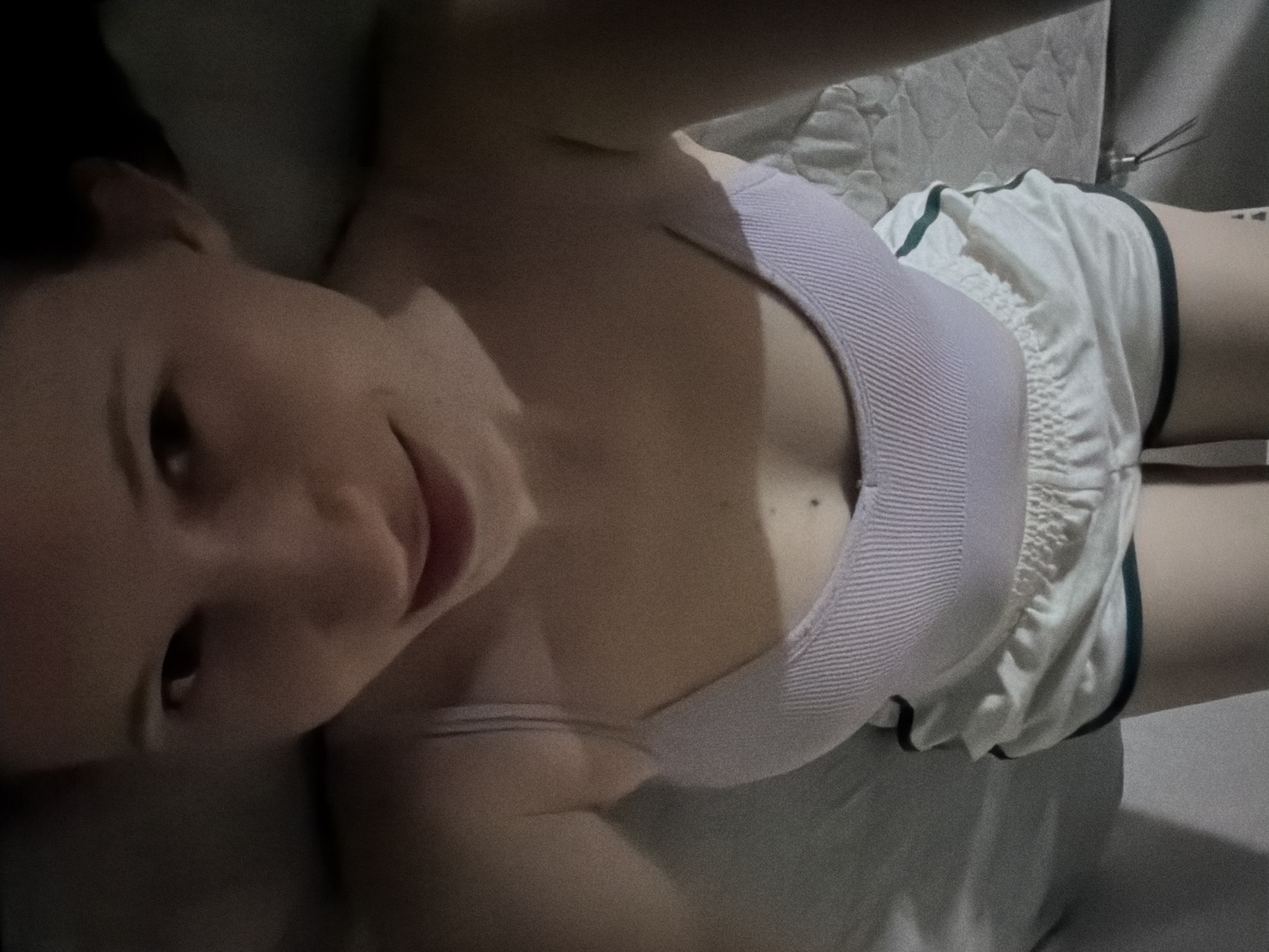 A woman lies on her back on a bed, looking up at the camera. She is wearing a light purple ribbed tank top and white shorts with dark green trim. Her left arm is bent and resting on her head. Her expression is relaxed and she appears to be smiling slightly. The lighting is dim, suggesting it is nighttime or the room is not well-lit. The background shows a quilted pillow and the edge of a mattress. There is a tattoo visible on her right hip. There is no visible text in the image.